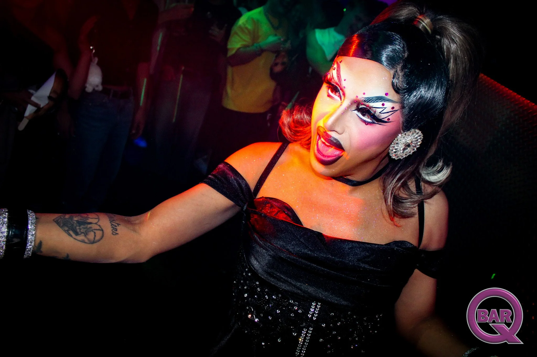 A drag queen performer with dramatic makeup, black and red hair, and rhinestone earrings singing into a microphone in a dimly lit bar or club.