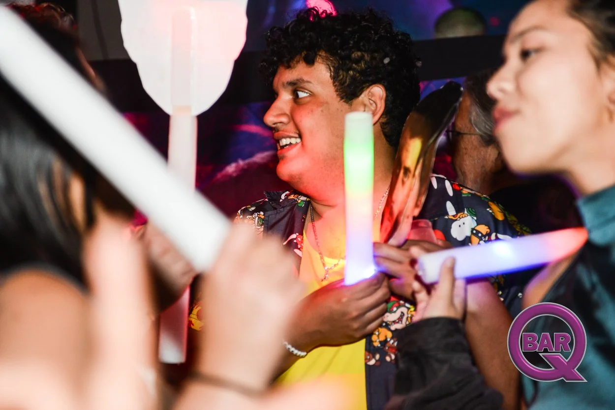 People holding light-up glow sticks at a lively party or nightclub, with colorful lighting and a logo that says 'Bar Q' in the bottom right corner.