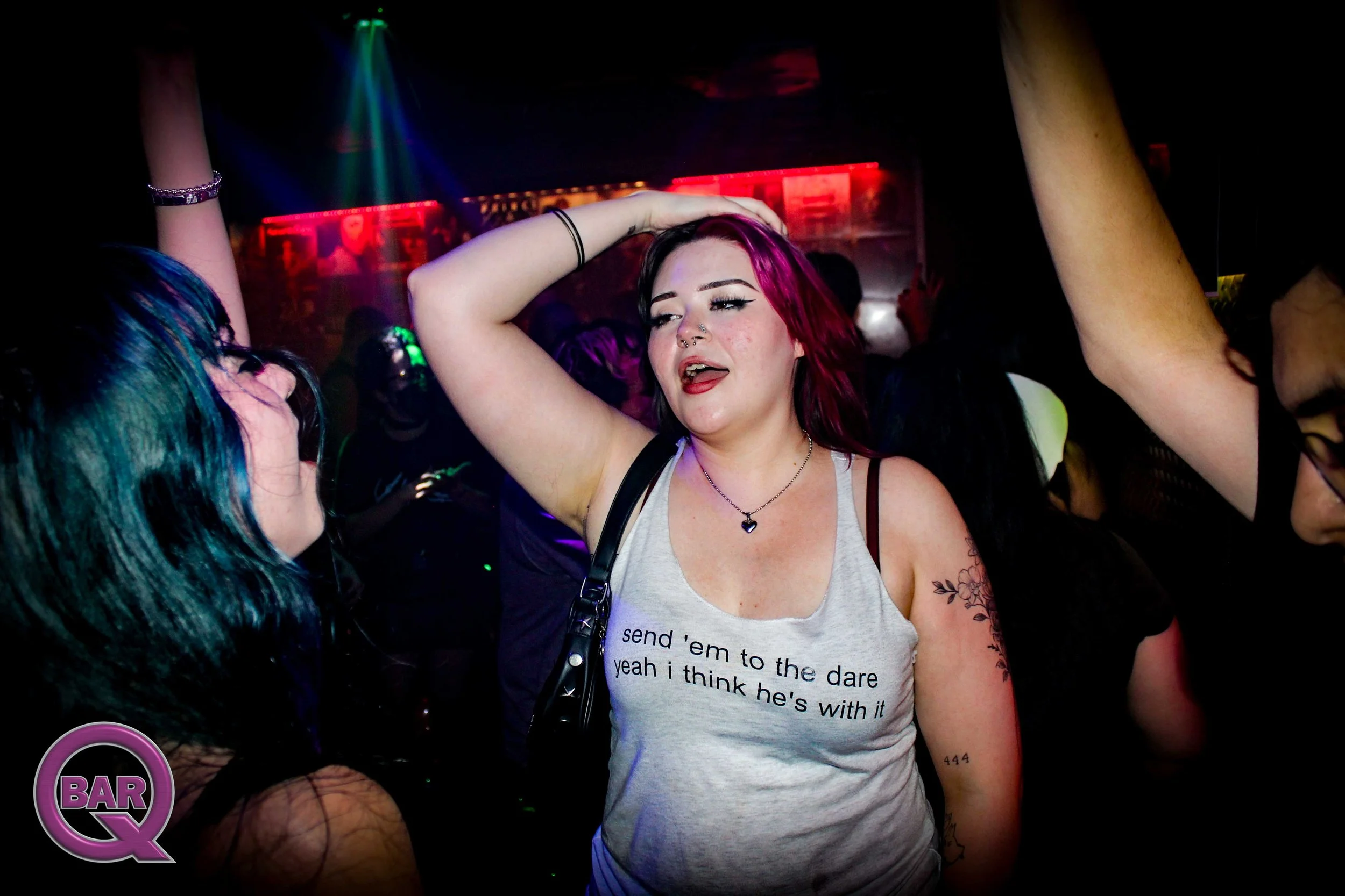 A woman with pink and black hair dancing in a dark nightclub, wearing a gray tank top with a read and black design, surrounded by other people, colorful lights overhead.