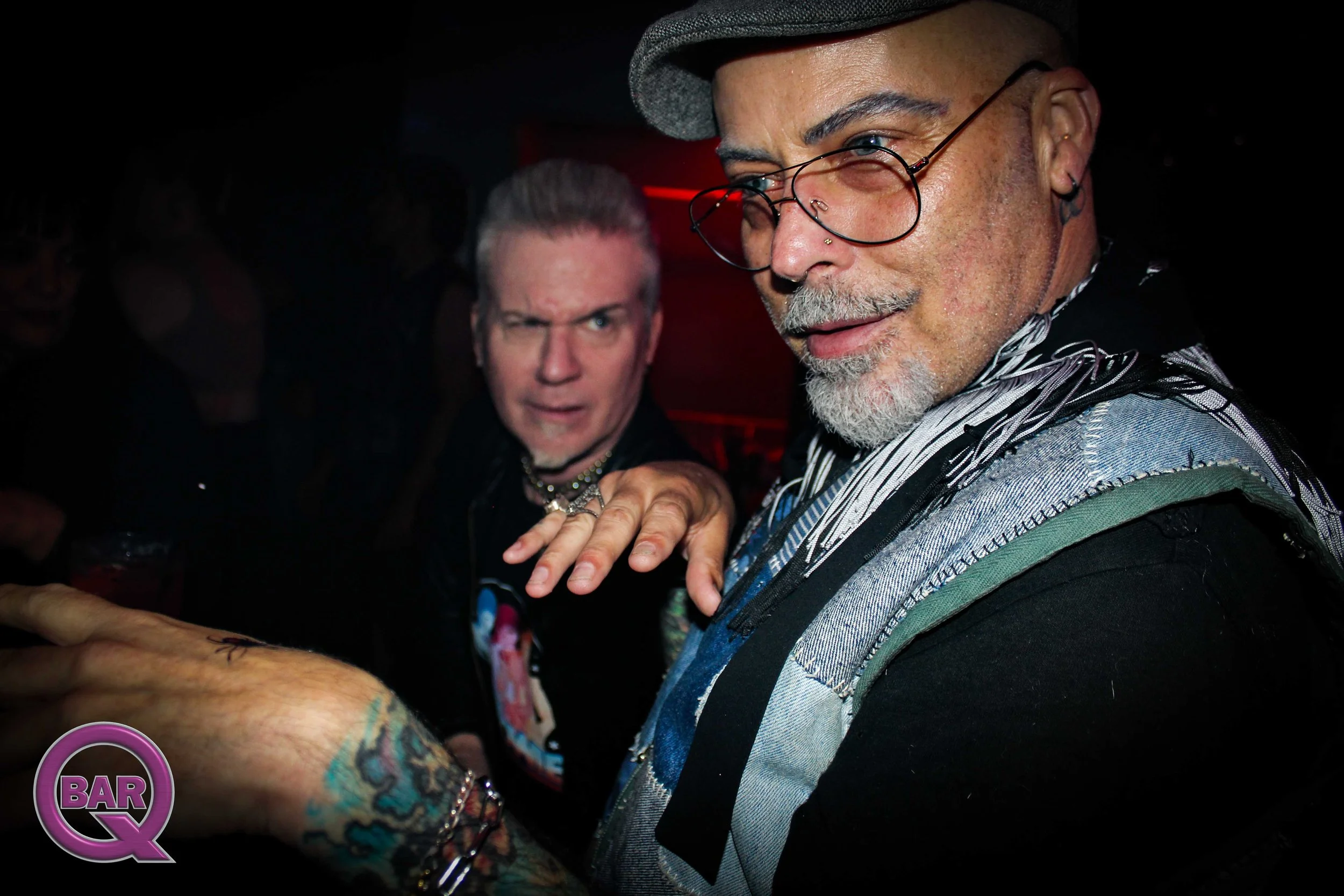 A close-up of a man with tattoos on his arms, wearing glasses, a hat, and a striped shirt, showing his hand and arm with tattoos, and another man with short hair and tattoos in the background.