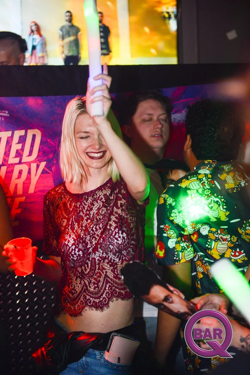 A woman in a lace top dancing with a smile on her face, holding a green glow stick, at a lively nightclub. She has blonde hair, dark lipstick, and a smartphone tucked into her waistband. The background features other partygoers, colorful lighting, an