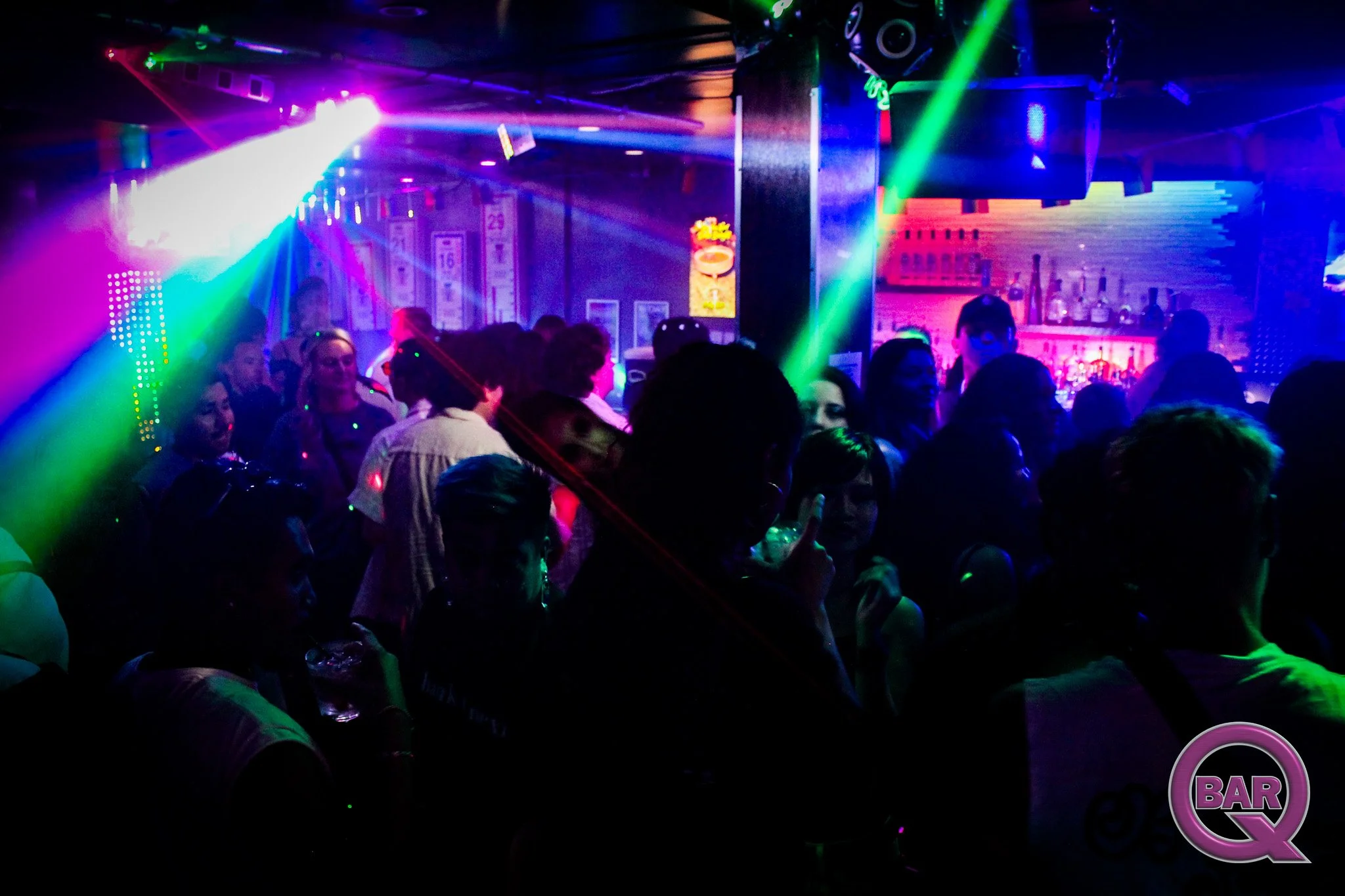 A crowded nightclub with vibrant colorful lighting, people dancing and socializing, a bar in the background with drinks, and a neon 'BAR' sign.