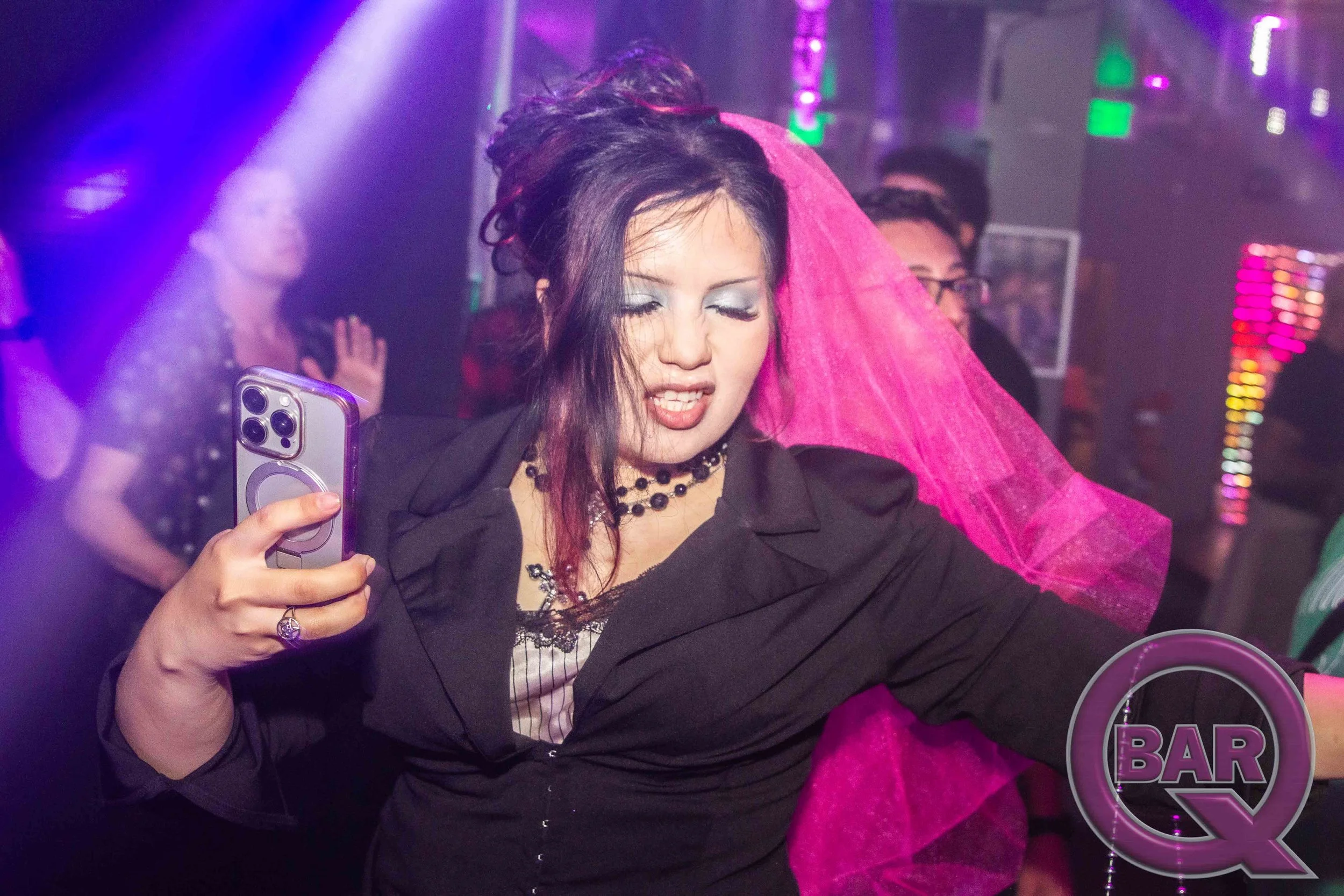 A woman with dark hair and makeup, wearing a black jacket and necklace, taking a selfie at a bar or nightclub, with purple lighting and neon signs in the background.