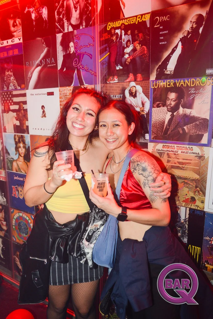 Two women smiling and holding drinks at a bar, standing in front of a wall covered with 1980s and 1990s music album covers.