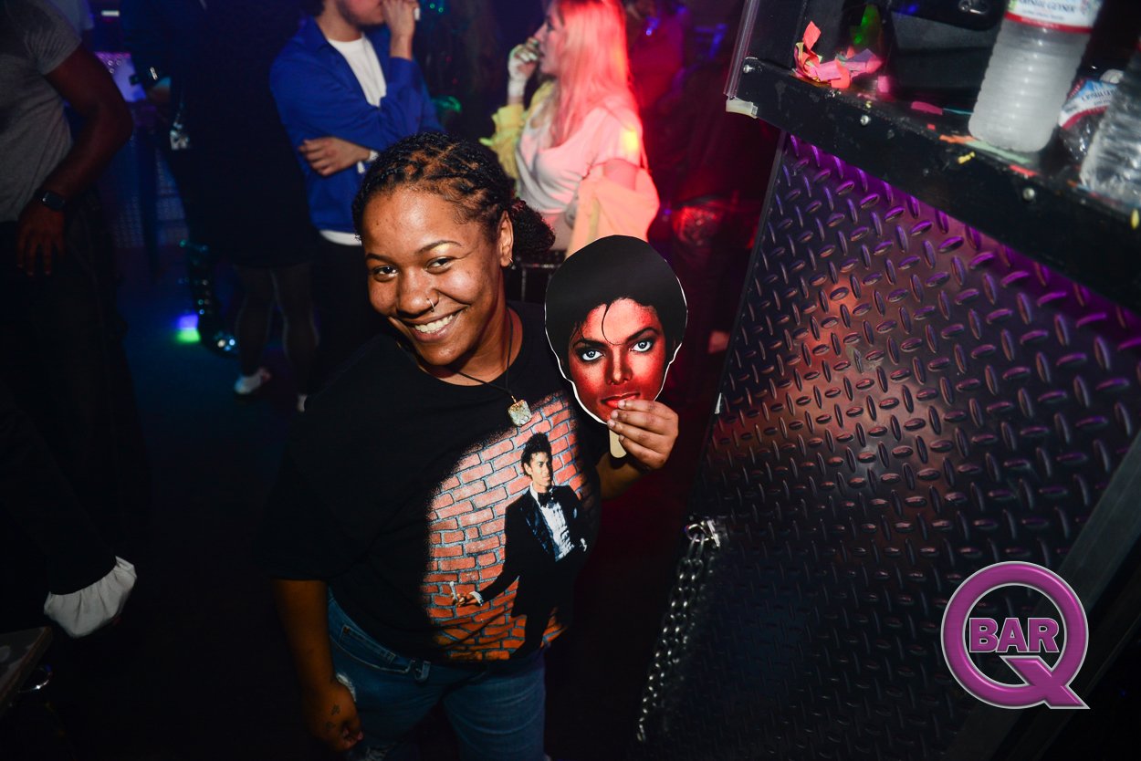 A smiling woman holding a cutout of a woman's face at a nightclub with other people in the background and purple and red lighting.