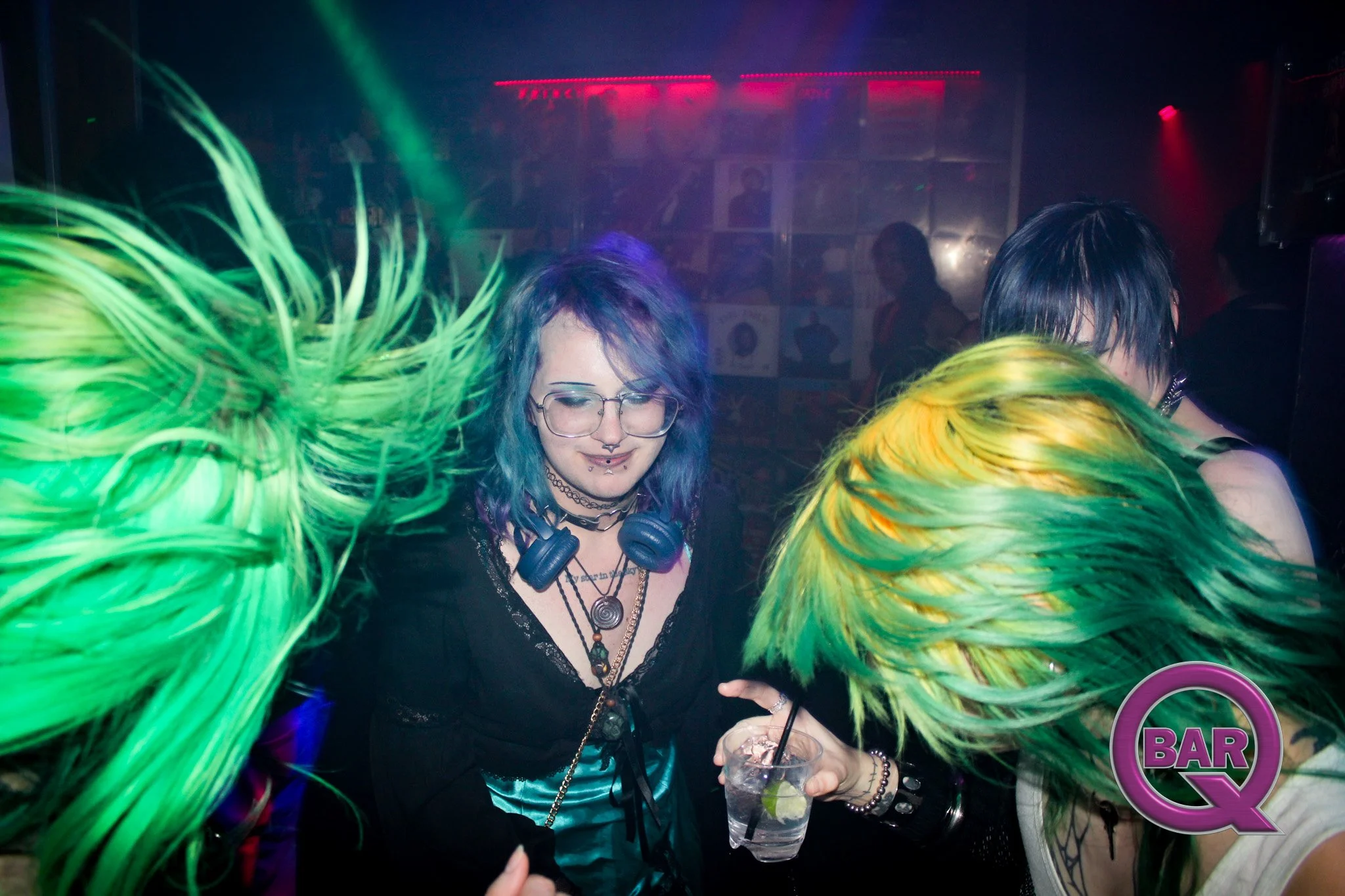 People with colorful hair dancing at a nightclub with green and red lights, drinking cocktails.