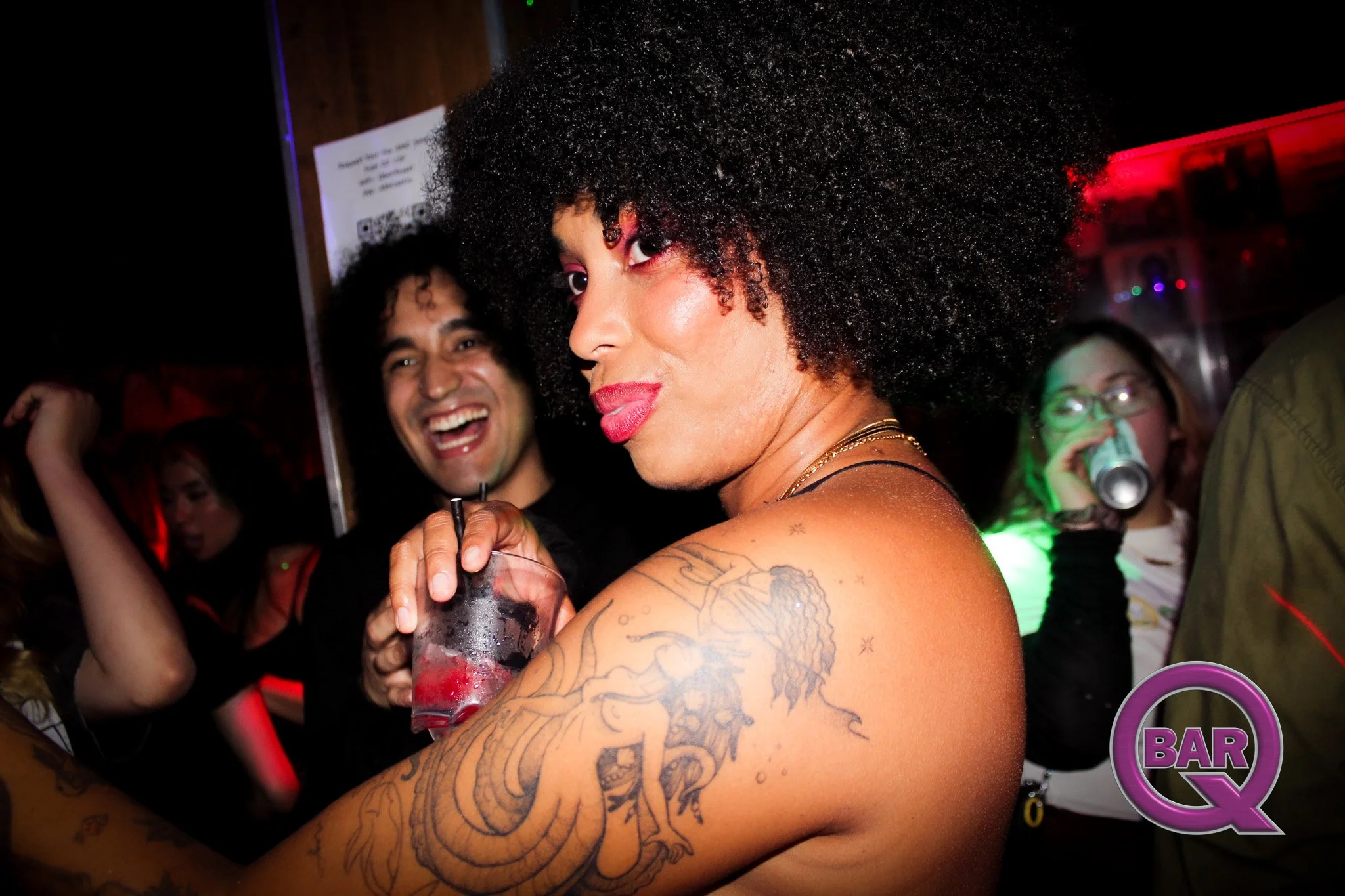 A woman with curly hair, visible tattoos, and makeup, holding a drink, in a lively bar or club with people smiling and drinking in the background.