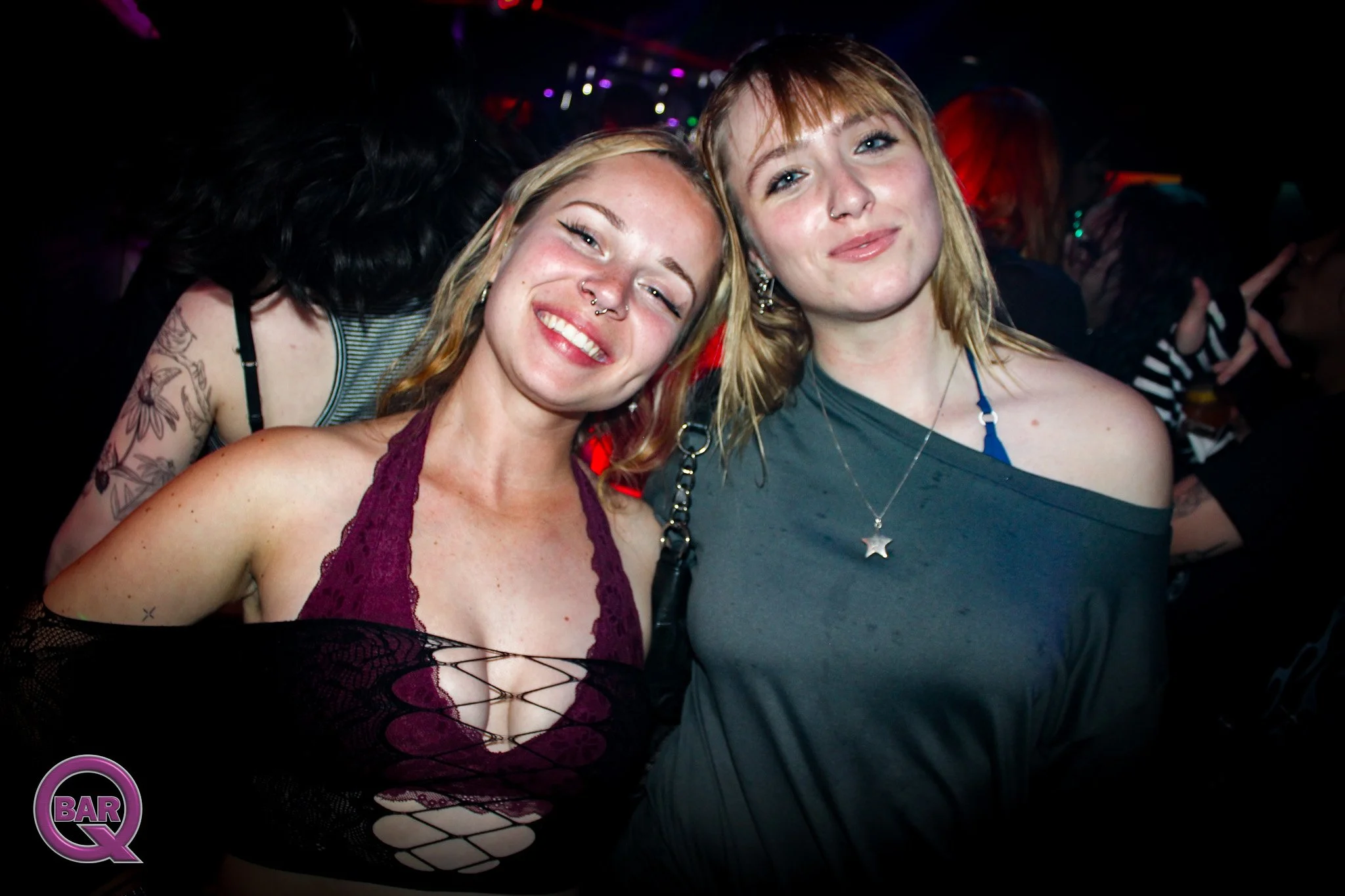 Two women smiling at a nightclub dance floor, with colorful lights and other people in the background.