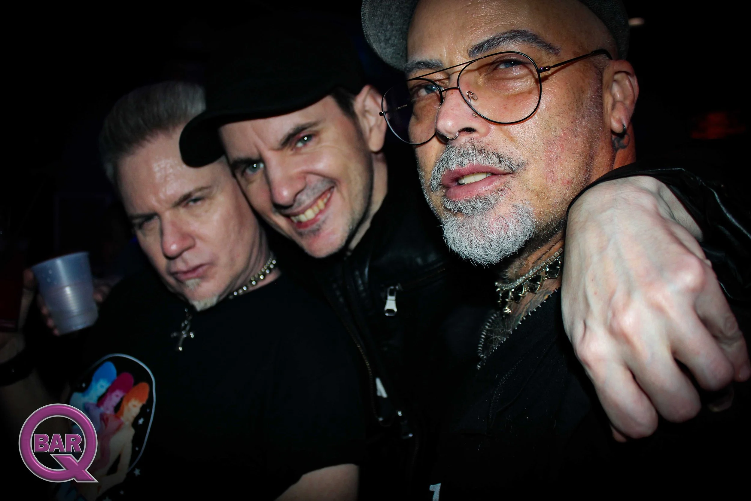 Three men at a bar, close-up. The man on the right has glasses, a gray beard, and earrings. The man in the middle has dark hair and is smiling. The man on the left has a serious expression, short hair, and is holding a drink.