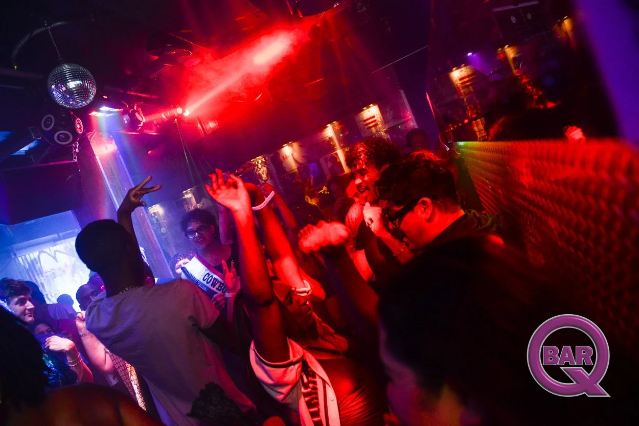 People dancing and enjoying themselves at a nightclub with colorful lights, a disco ball, and a bar logo in the corner.