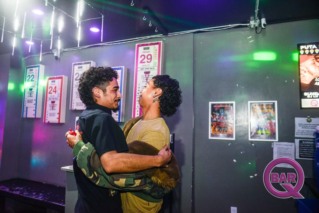 A young man and woman are dancing closely together in a nightclub or bar, with colorful lights and posters on the wall behind them.