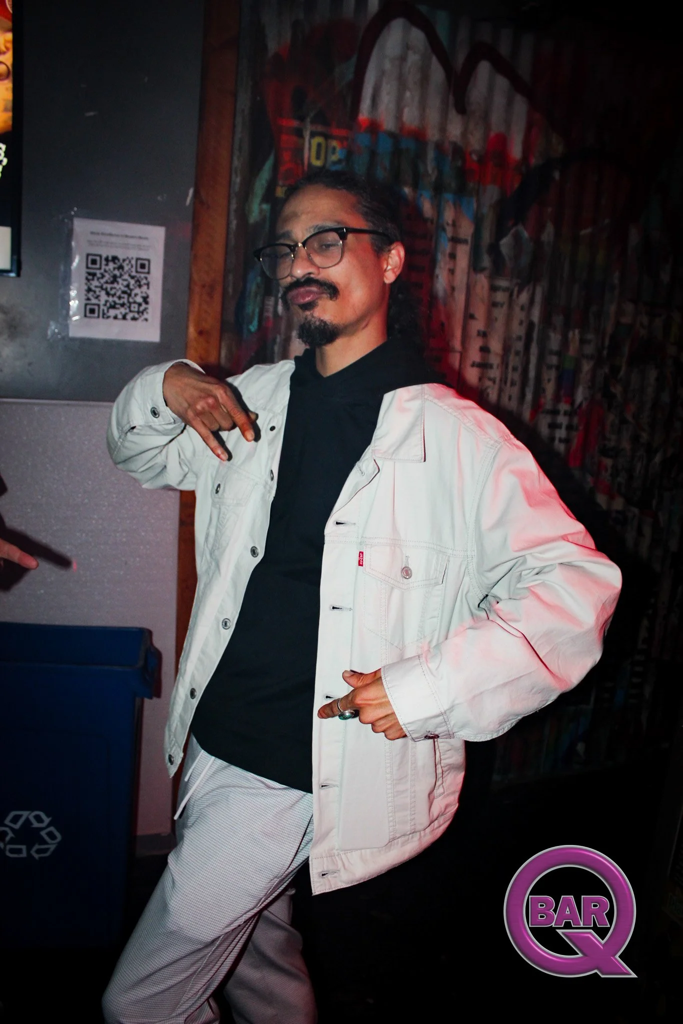 A man with glasses and facial hair, wearing a white jacket and black shirt, making a peace sign with his right hand in a bar setting with colorful graffiti on the wall behind him.