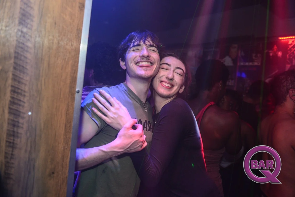 A young man and woman hugging and smiling in a bar or nightclub with purple and red lighting, and people in the background.