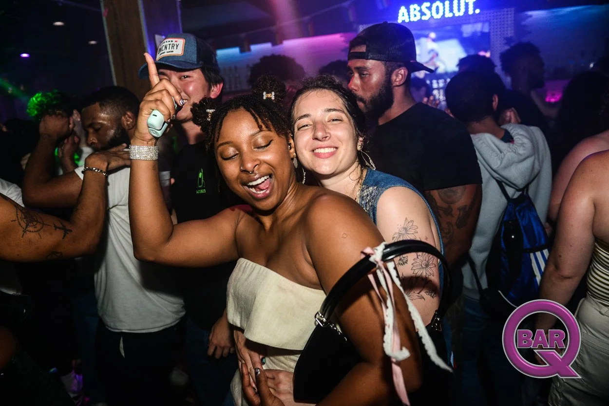 Two women dancing and smiling at a nightclub with colorful lights and a crowd of people in the background.