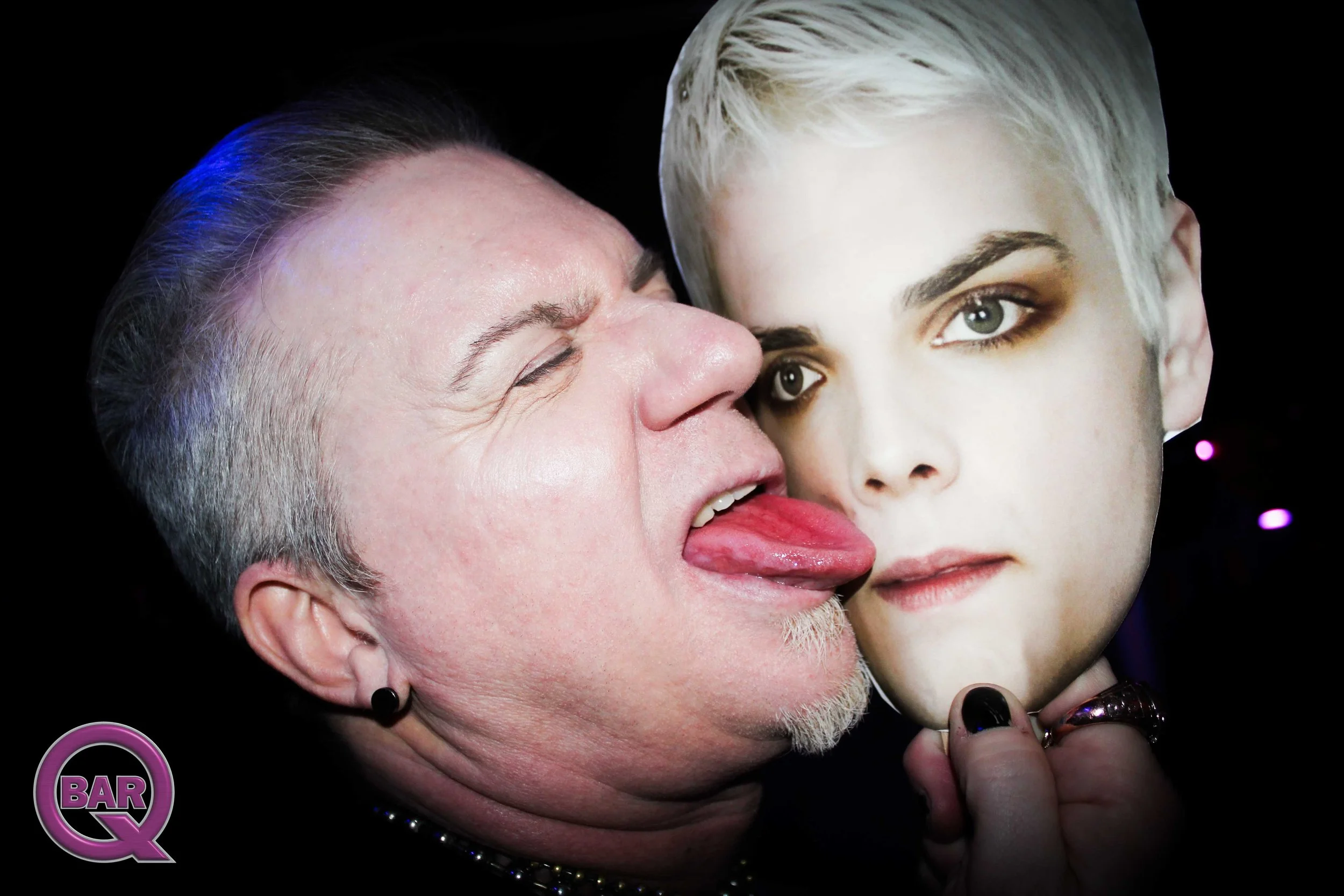 A man with gray hair and a woman with blonde hair and green eyes are close together, with the man playfully sticking out his tongue near the woman's face against a black background.