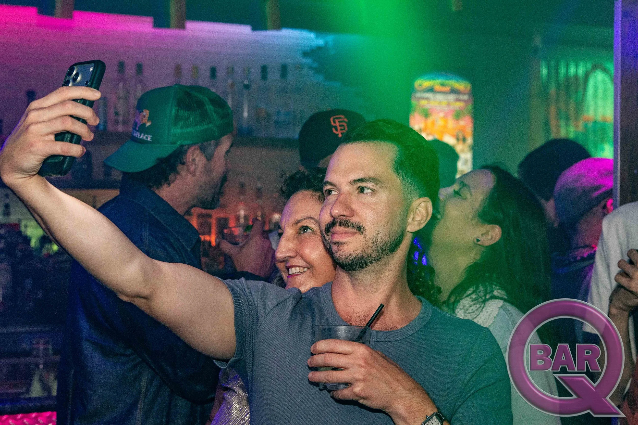 Two people taking a selfie at a lively bar or club with colorful neon and LED lighting, and a crowd in the background.