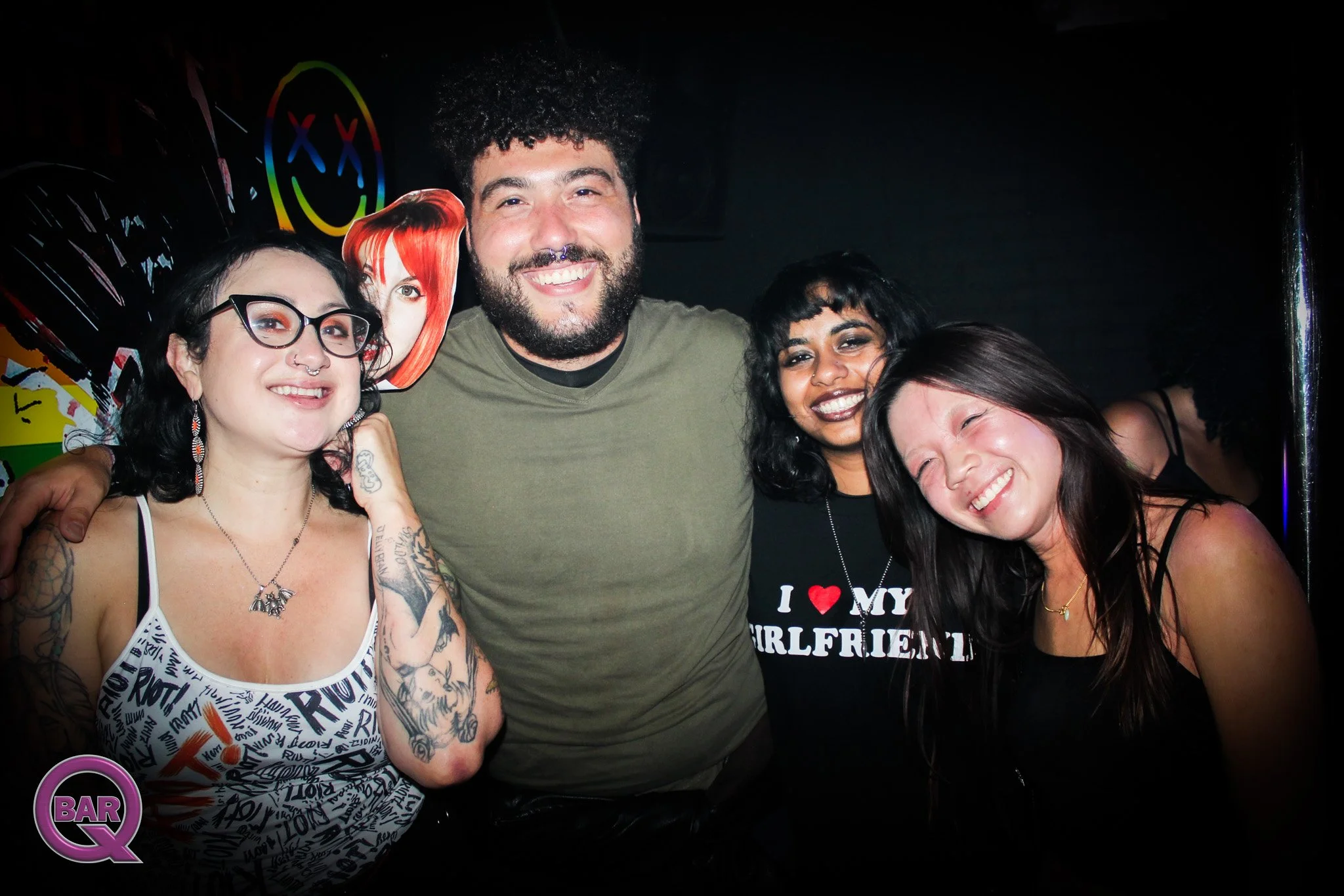 Group of five diverse friends smiling and posing together at a bar or nightclub, with dark background and colorful neon lighting.