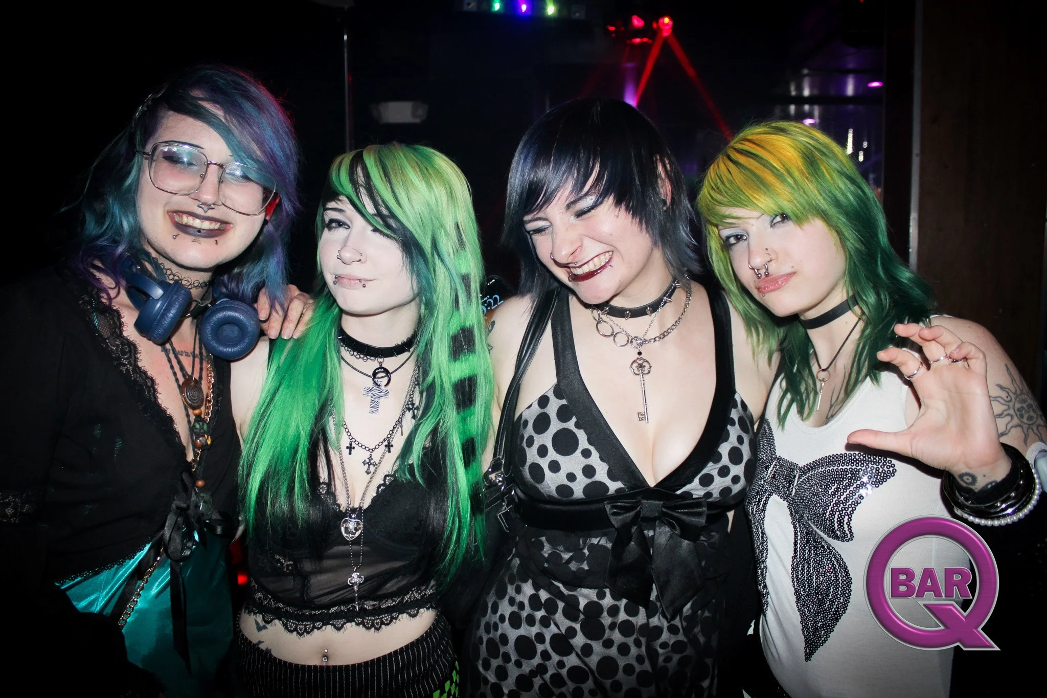 Four women with brightly colored hair and piercings smiling and posing together in a nightclub with neon lights in the background.