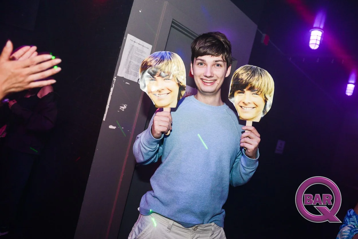 A young man in a blue sweater smiling and holding two masks of a boy's face, in a dimly lit bar or nightclub with purple lights.