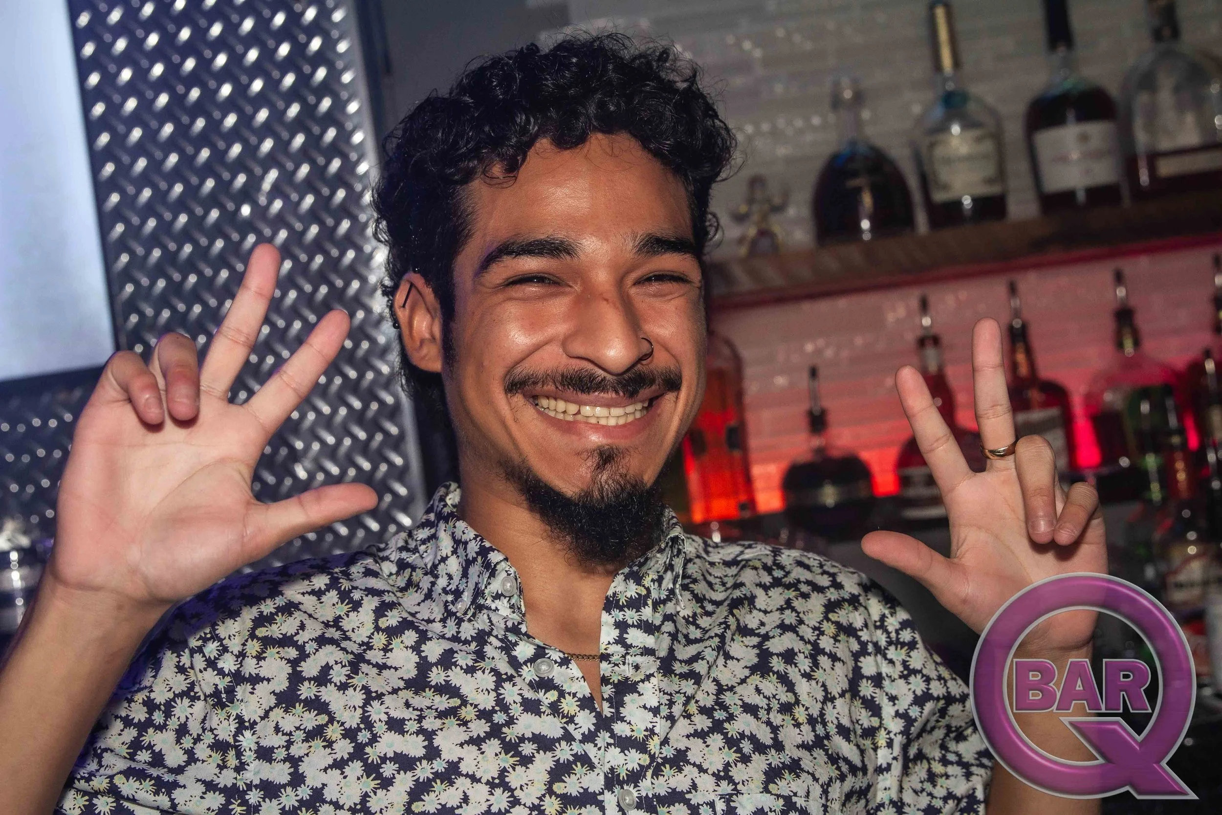 A man with curly black hair and a mustache, smiling and making peace signs with both hands. He is wearing a patterned shirt and standing in a bar setting with bottles and bar equipment in the background.