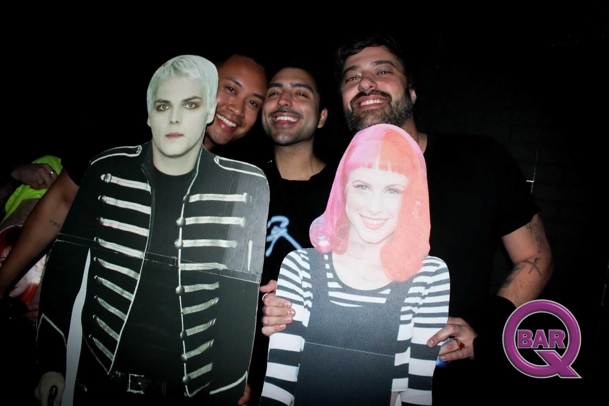A group of four people holding cardboard cutouts with the faces of two women and two men, smiling and posing together, with a dark background and a purple and black circular logo with the word "BAR" in the bottom right corner.