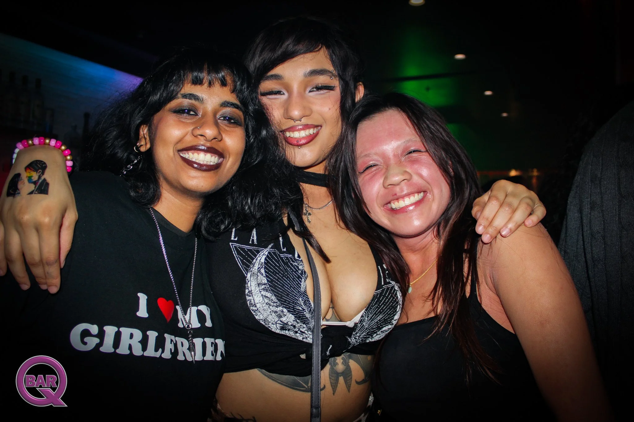 Three women smiling and posing together at a bar or club, with dark background and colorful lighting.