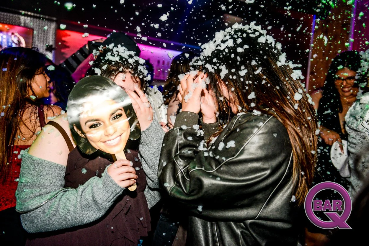 People celebrating at a foam party in a nightclub, with foam falling from above and a woman smiling holding a cardboard cutout mask of a woman's face. The scene is vibrant with colorful lighting.