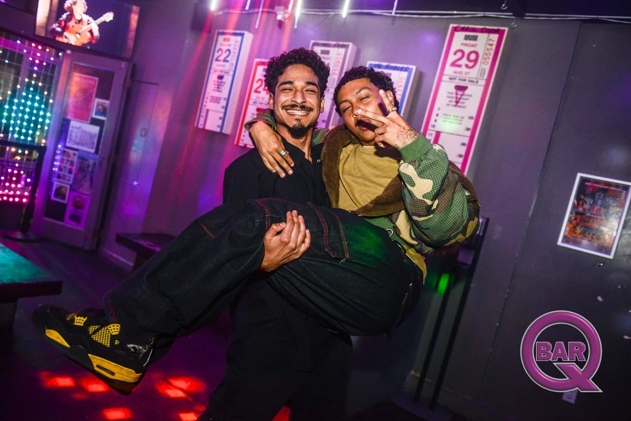 Two smiling men are celebrating and lifting a person in a bar with neon lights and posters on the wall.