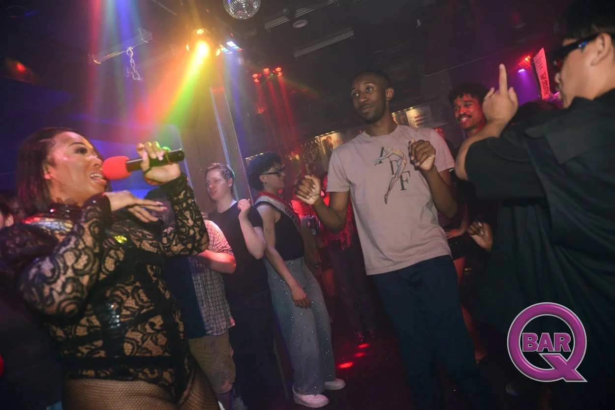 A woman singing into a red microphone on a dimly lit dance floor with colorful lights, while a group of people dance and socialize around her at a bar or nightclub.