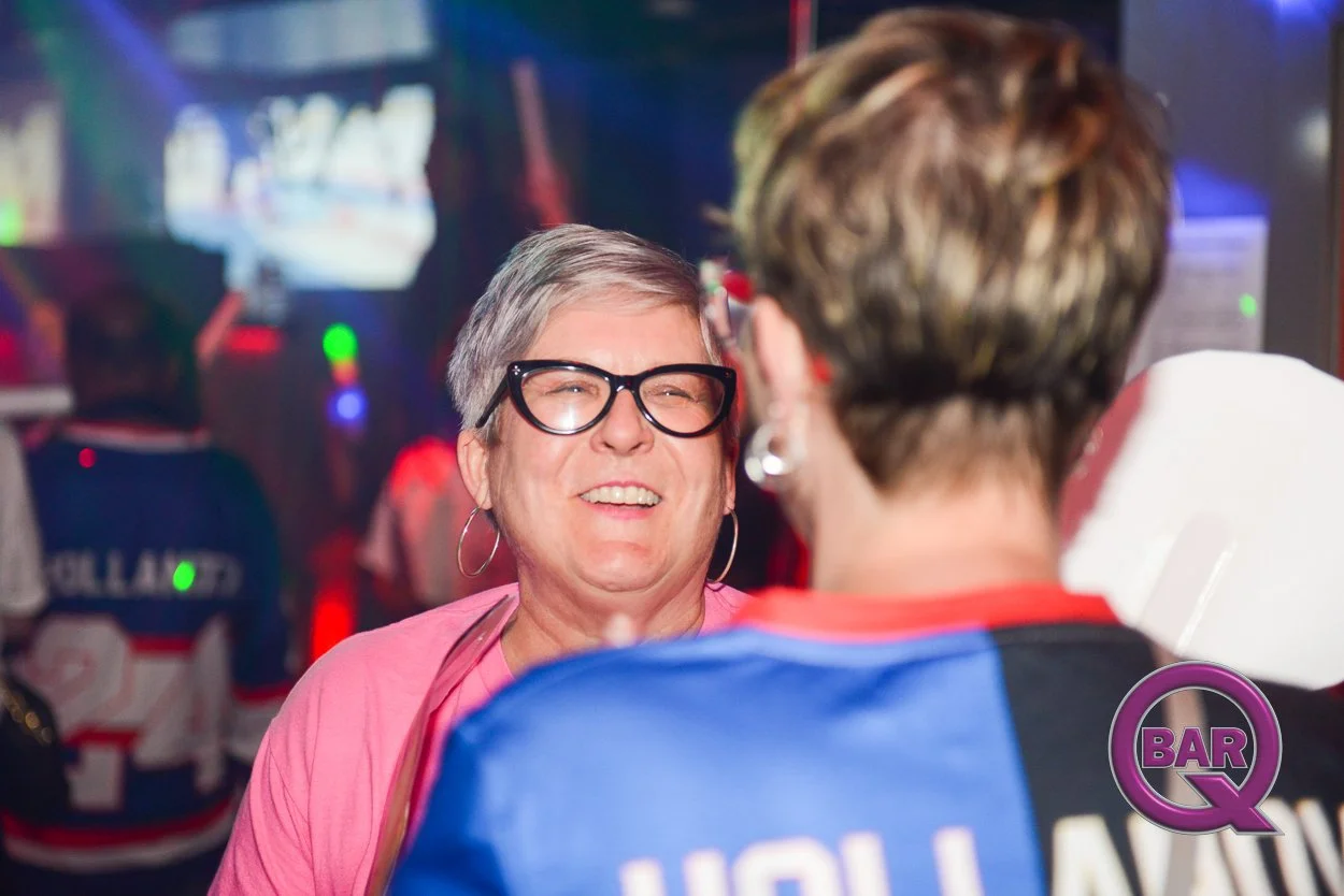 Two women engaged in conversation at a sports bar, with screens displaying a hockey game in the background. One woman has short gray hair, glasses, and hoop earrings, wearing a pink top. The other has short brown hair. The bar logo 'BAR Q' is visible