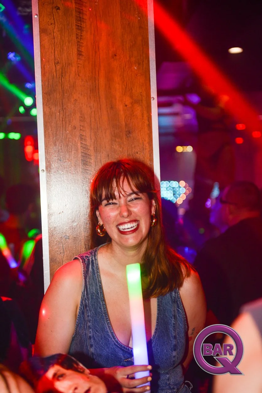 A woman with shoulder-length brown hair, wearing a sleeveless denim top, smiling and holding a glowing rainbow-colored light stick at a lively nightclub or bar, with colorful lights and blurred crowd in the background.