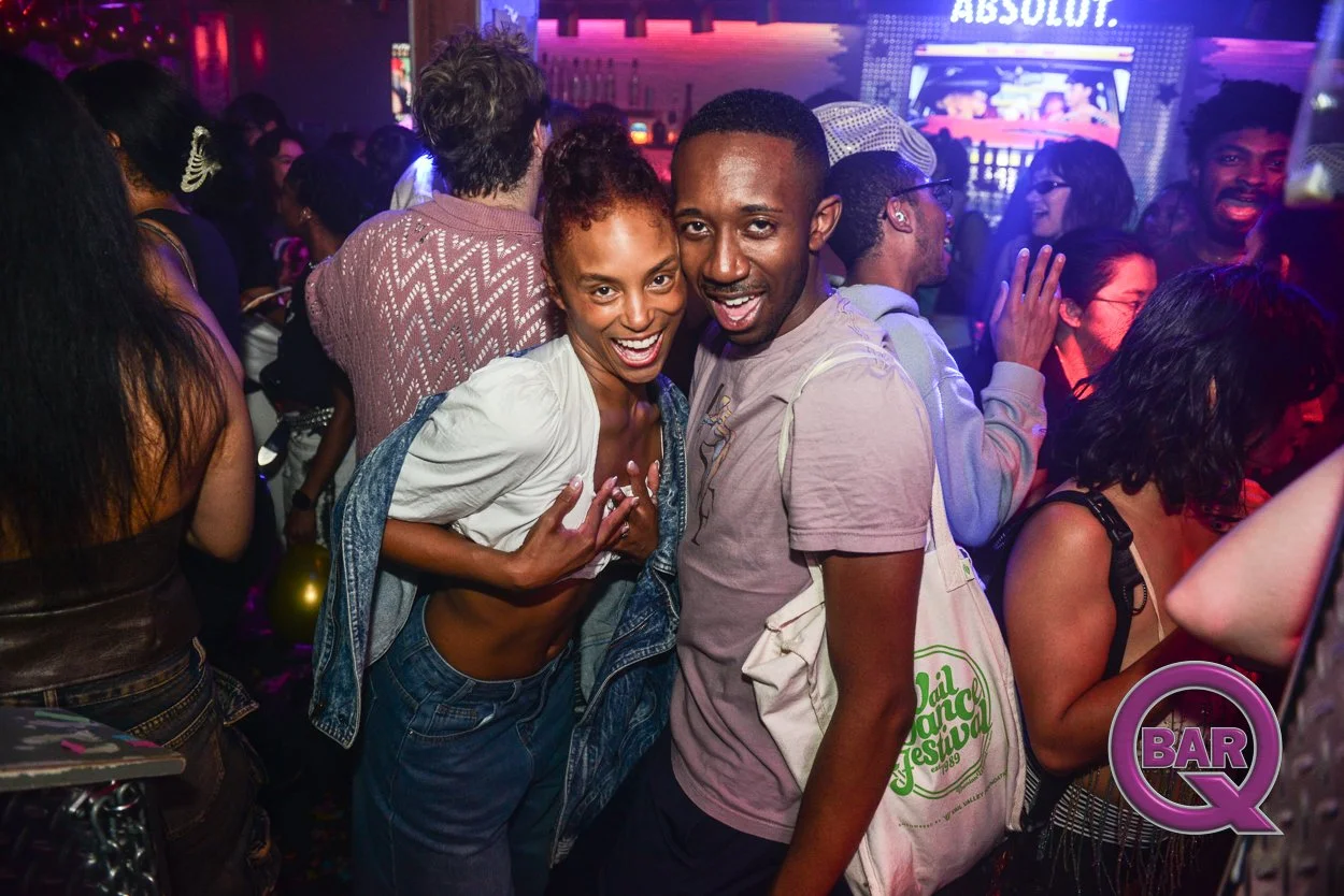 Two smiling young adults, a woman and a man, pose together at a lively nightclub. The woman has curly hair and wears a denim shirt with a yellow top underneath, while the man has short hair and wears a pink T-shirt, holding a tote bag. The background