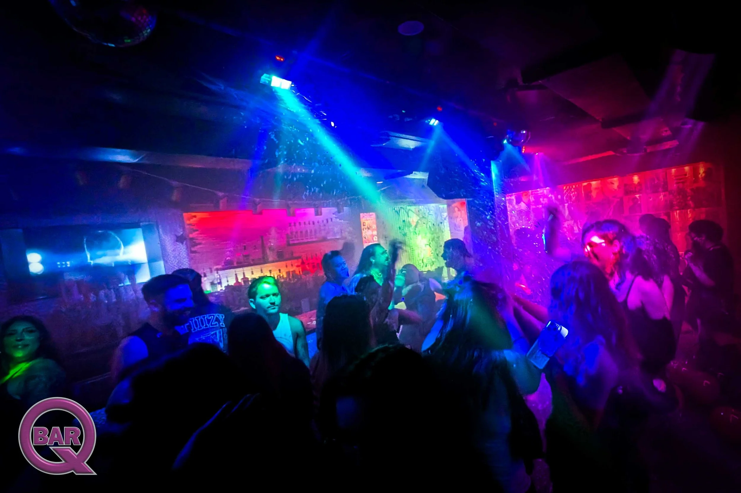 People dancing and socializing in a nightclub with colorful, vibrant lighting and a bar in the background.