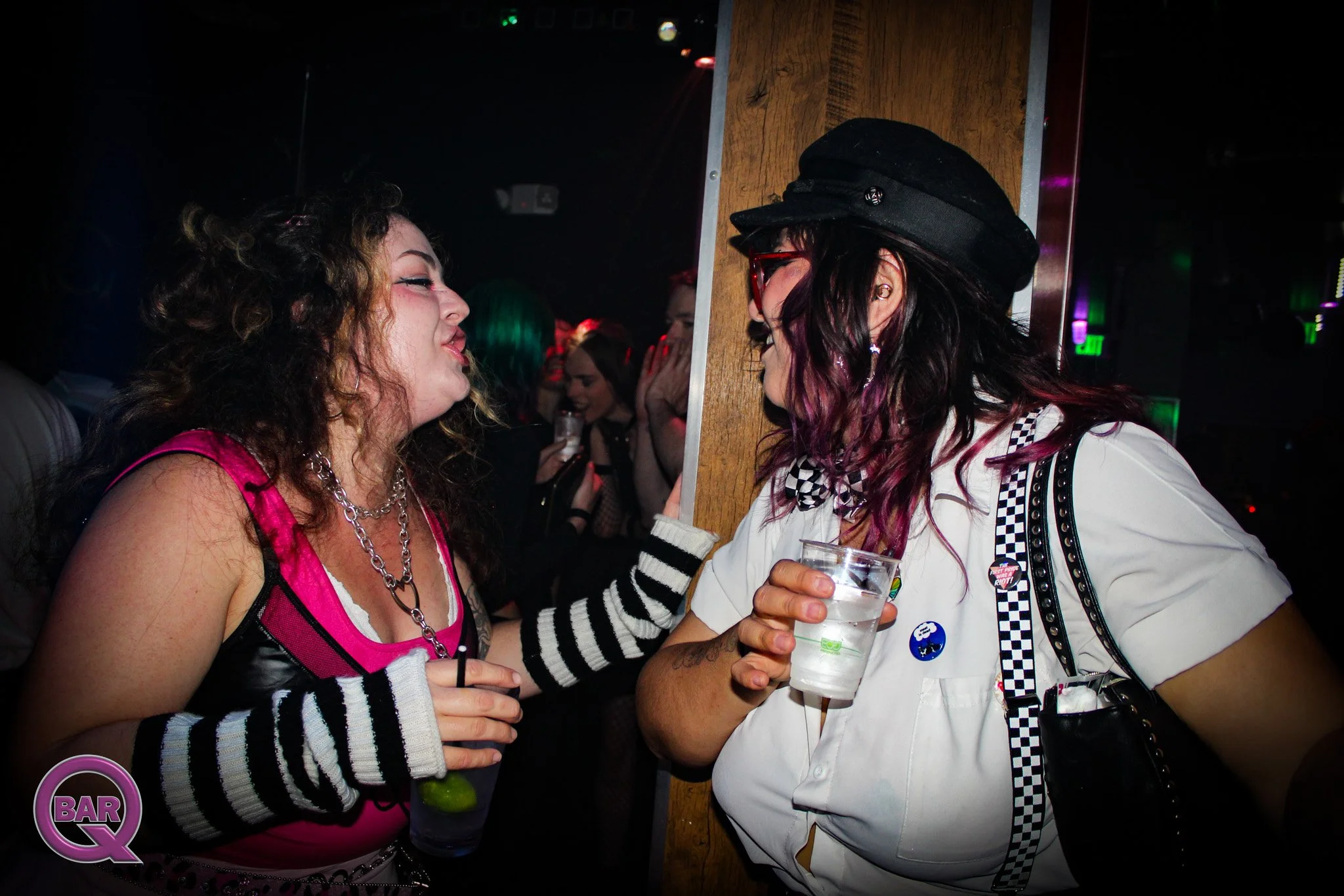 Two women talking closely in a bar or nightclub, one with curly hair and pink clothing, the other with long dark hair wearing a police-style uniform and hat.