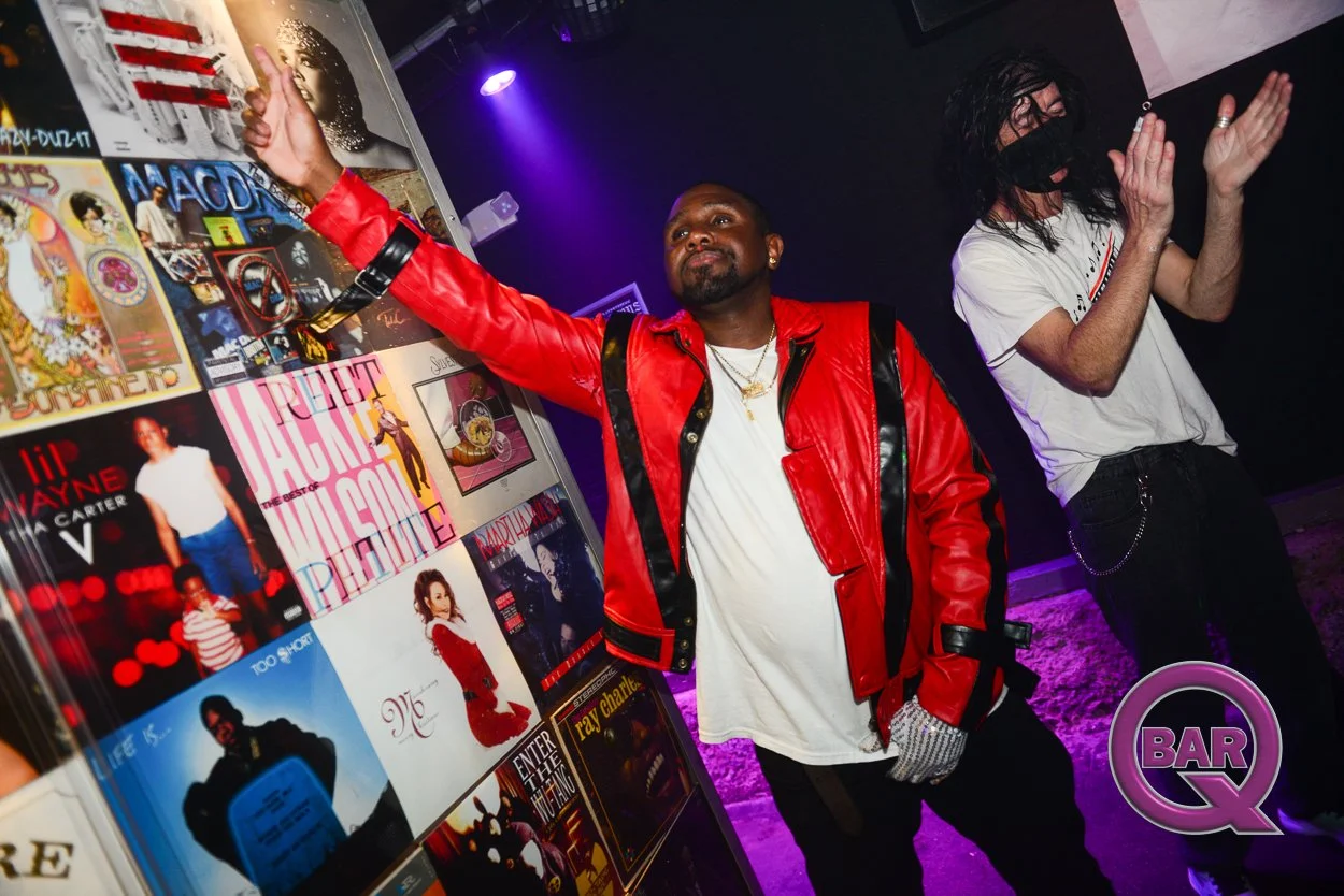 Two men in a nightclub, one wearing a red and black leather jacket and pointing at a wall with album covers, the other with long hair, wearing a white t-shirt and a black face mask.