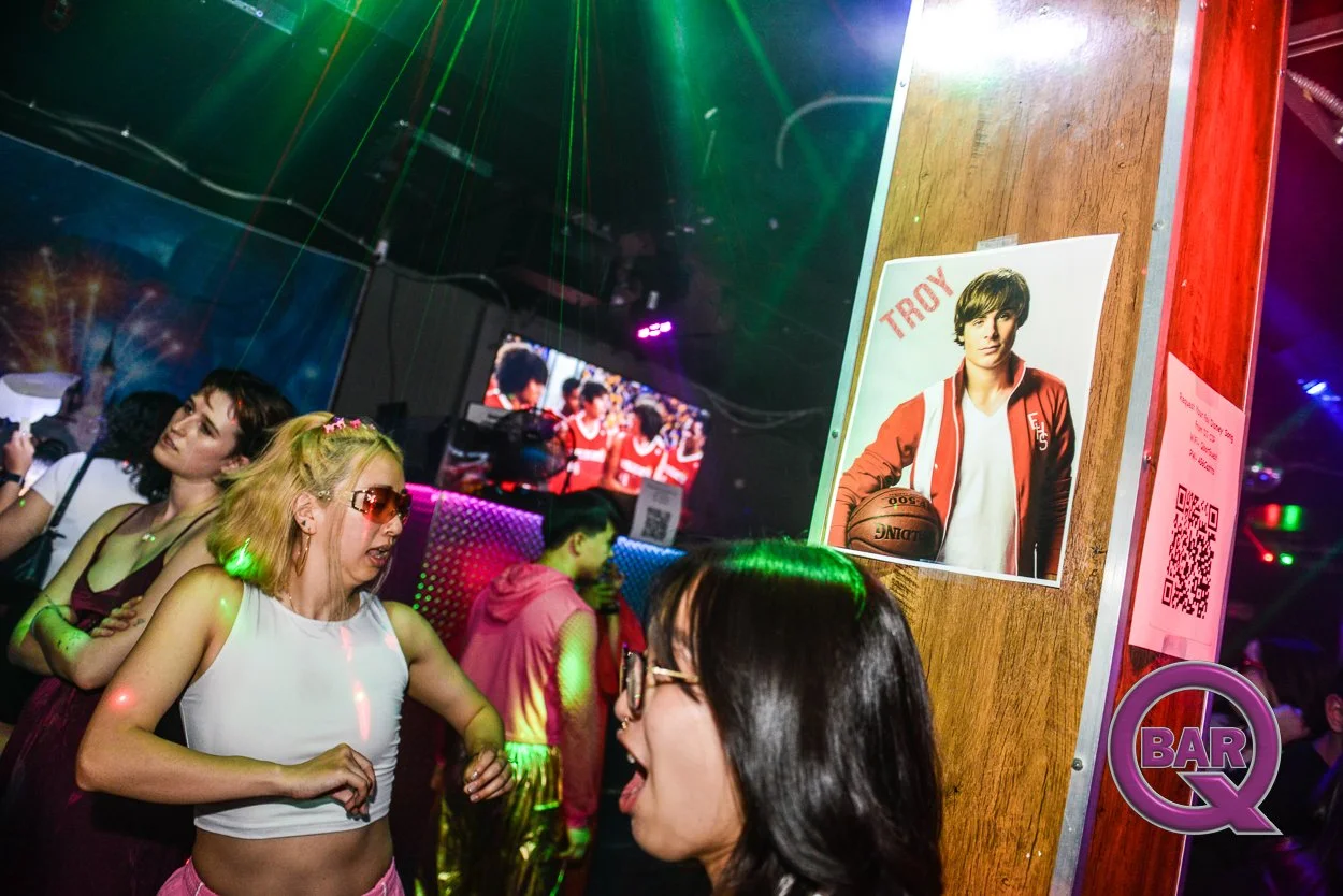 People dancing in a nightclub with colorful lights, a poster of a teenage boy holding a basketball, and multiple TV screens showing a group of young people in red uniforms.