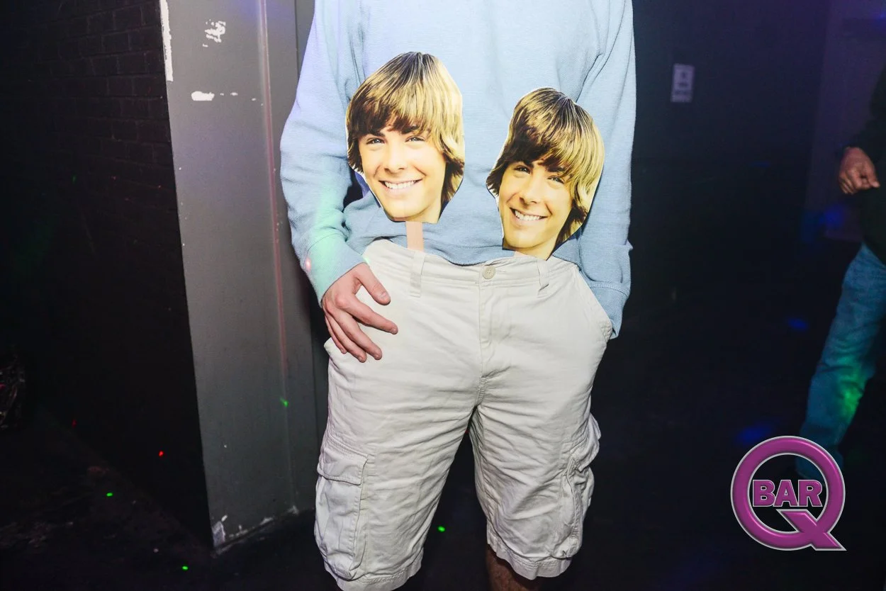 Person wearing a light blue hoodie and beige cargo shorts holding two cardboard cutouts of a smiling young man's face in front of their body at a nightclub or bar.