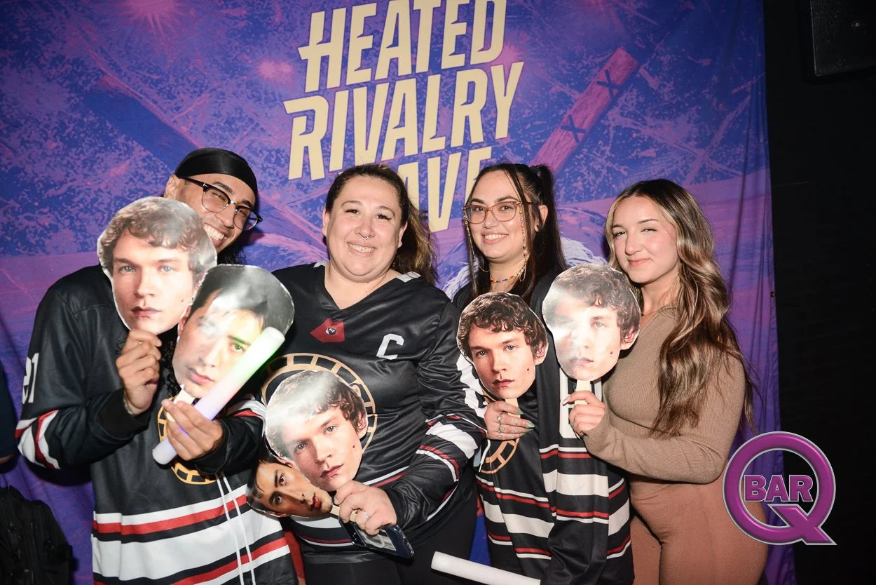 People posing indoors with fans of a TV show, holding cardboard cutouts of characters' faces, in front of a purple backdrop with the phrase 'Heated Rivalry' and 'Bar Q' logo.
