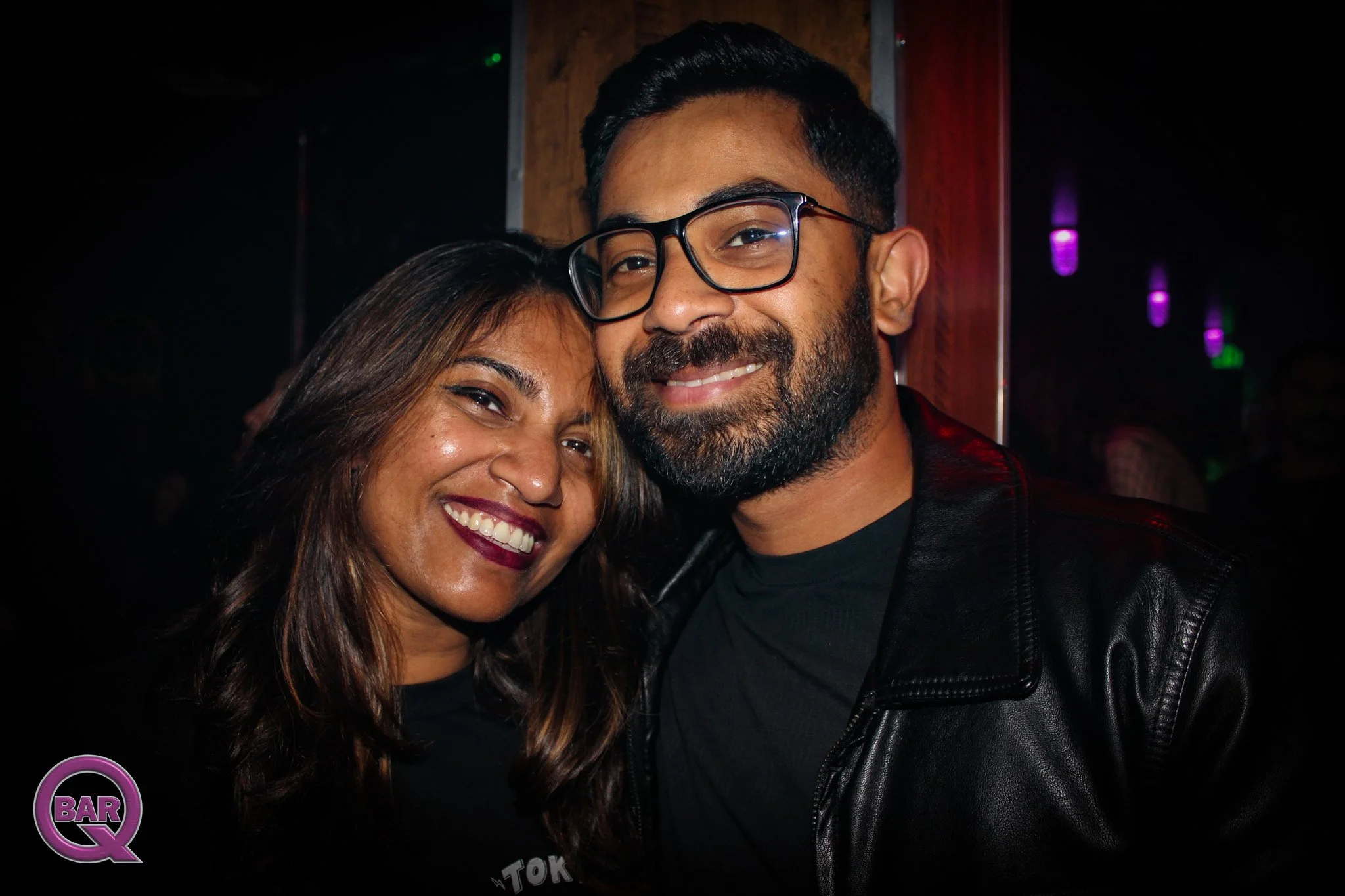 A smiling woman and man close together at a nightclub with purple and green lights, with a 'Q Bar' logo in the corner.