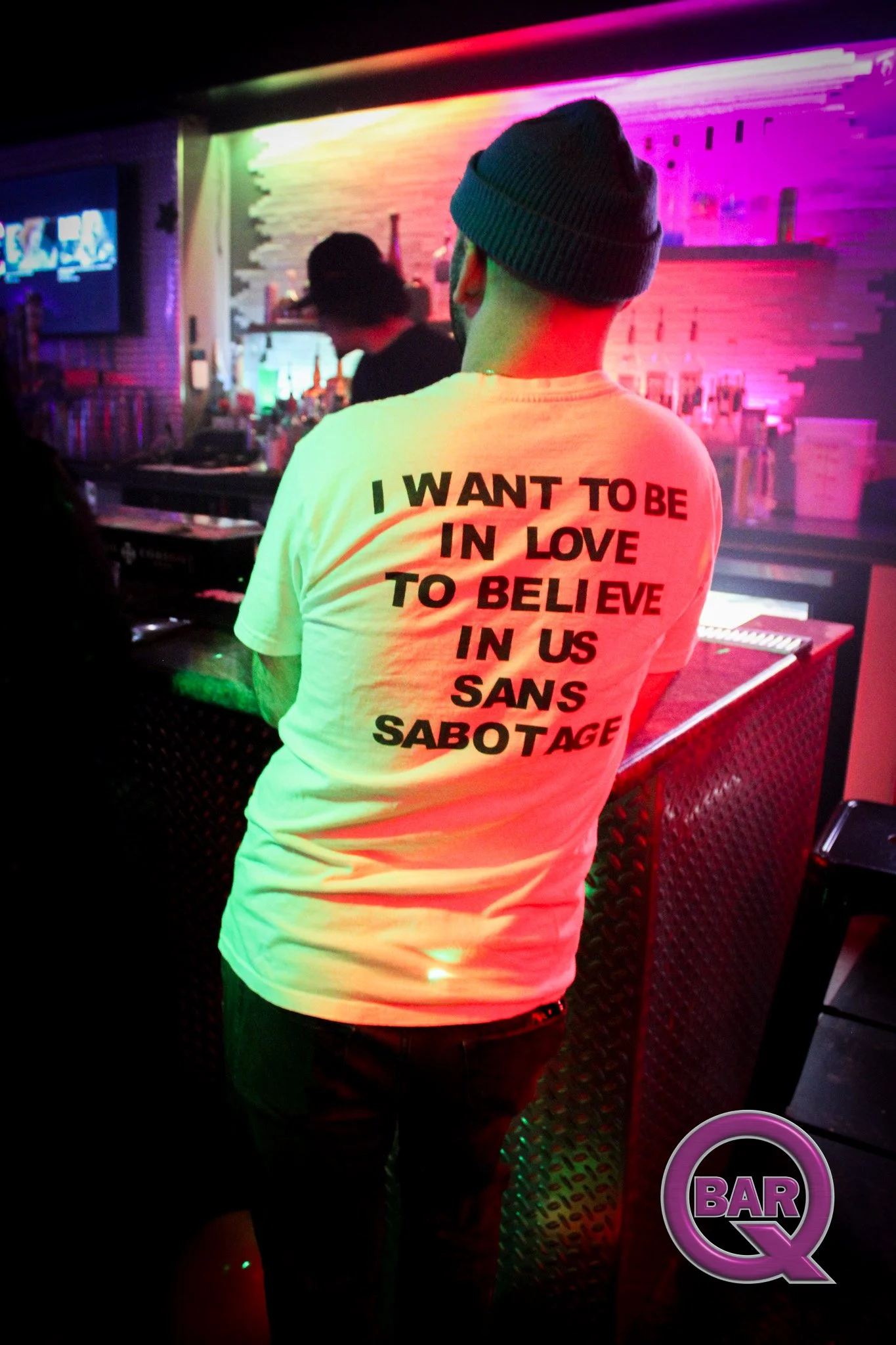 A person wearing a white T-shirt with black and red text that reads 'I WANT TO BE IN LOVE TO BELIEVE IN US SANS SABOTAGE' stands in a bar or nightclub setting. The individual has short hair and is wearing a gray beanie, with neon-colored lighting ill