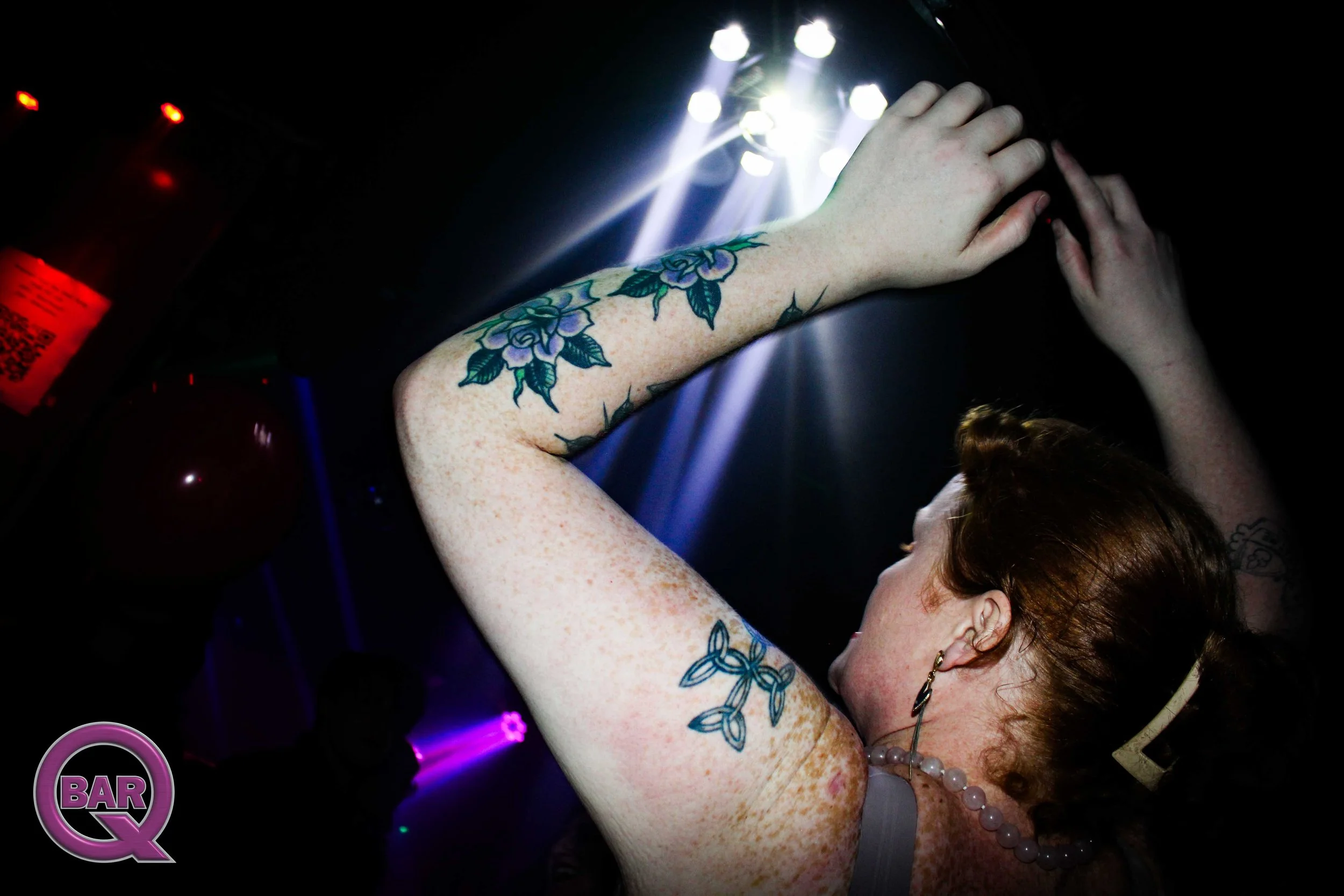 A woman with reddish-brown hair and tattoos on her arm is dancing or posing in a dark nightclub with bright stage lights overhead.