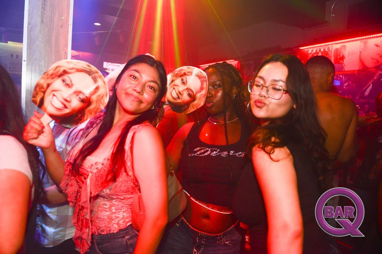Group of five women in a nightclub or bar with colorful lighting, holding paper masks with smiling faces, and a neon sign that says "BAR Q".