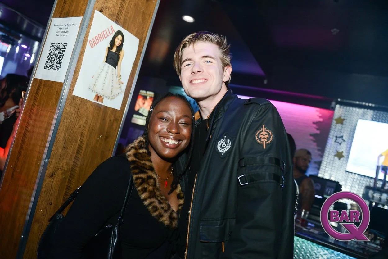 Two smiling people hugging at a nightclub, with a poster of a woman named Gabriella and a 'BAR Q' sign in the background.