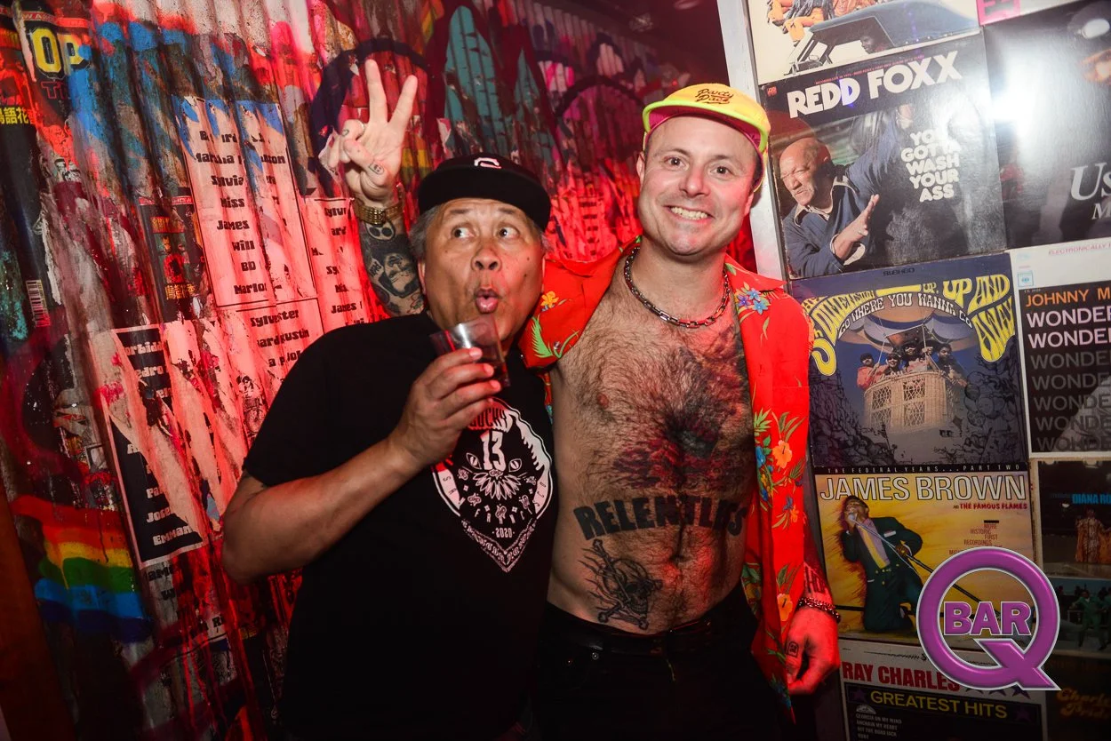 Two men at a music or retro themed party, one wearing a black T-shirt with a band logo and a cap, holding a drink, making a surprised face and peace sign, the other shirtless with chest hair, a vibrant jacket, and the word 'RELENTLESS' tattooed on hi
