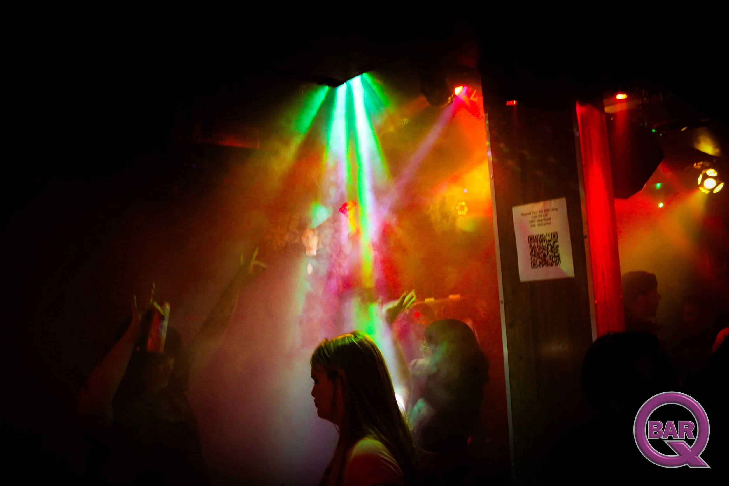 A dark scene in a nightclub with multicolored stage lights, including green, yellow, and red, illuminating the crowd and smoke. People are dancing and raising their hands.