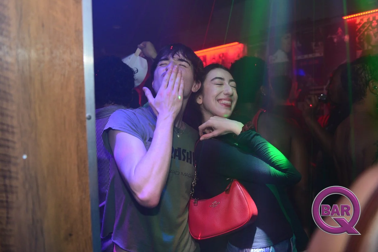 Two young people smiling and enjoying themselves at a nightclub or bar with colorful lights, surrounded by other people.