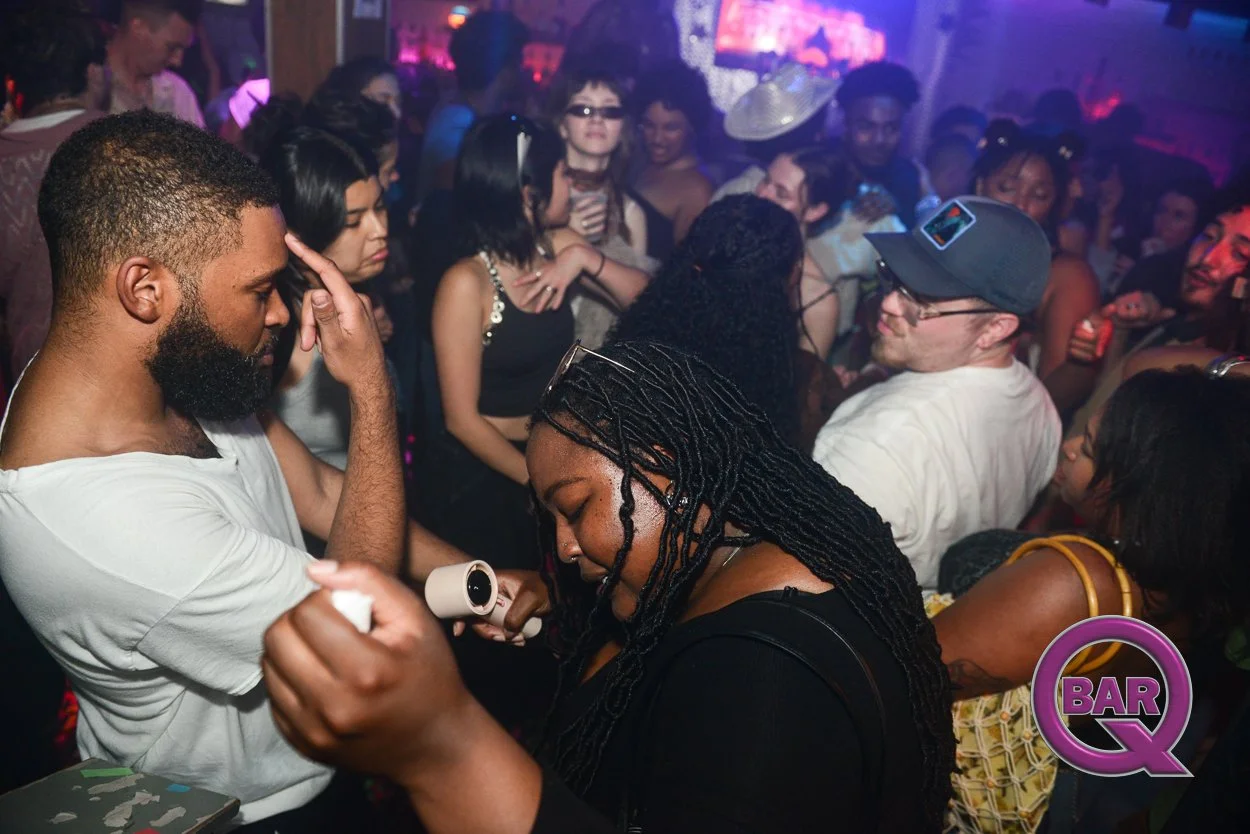 Crowd of people dancing and socializing at a nightclub with colorful lighting, including a woman with long braids in the foreground, a man with a beard wearing a white shirt, and a woman wearing a yellow dress, with a 'Bar Q' logo in the bottom right