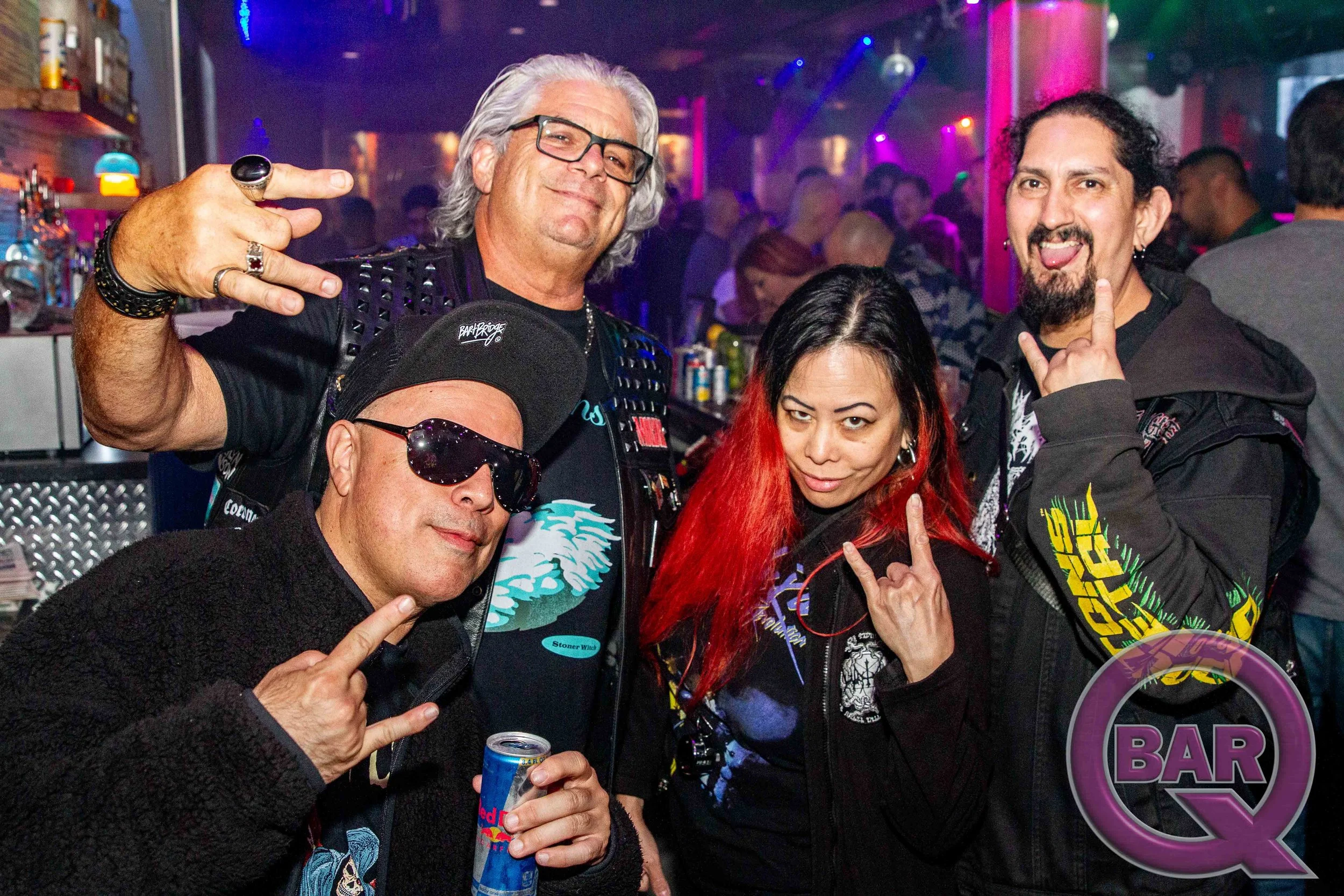 Group of five people at a bar or club enjoying themselves. They are making rock and roll hand signs, holding drinks, and smiling. The background has neon lights and a bar counter.