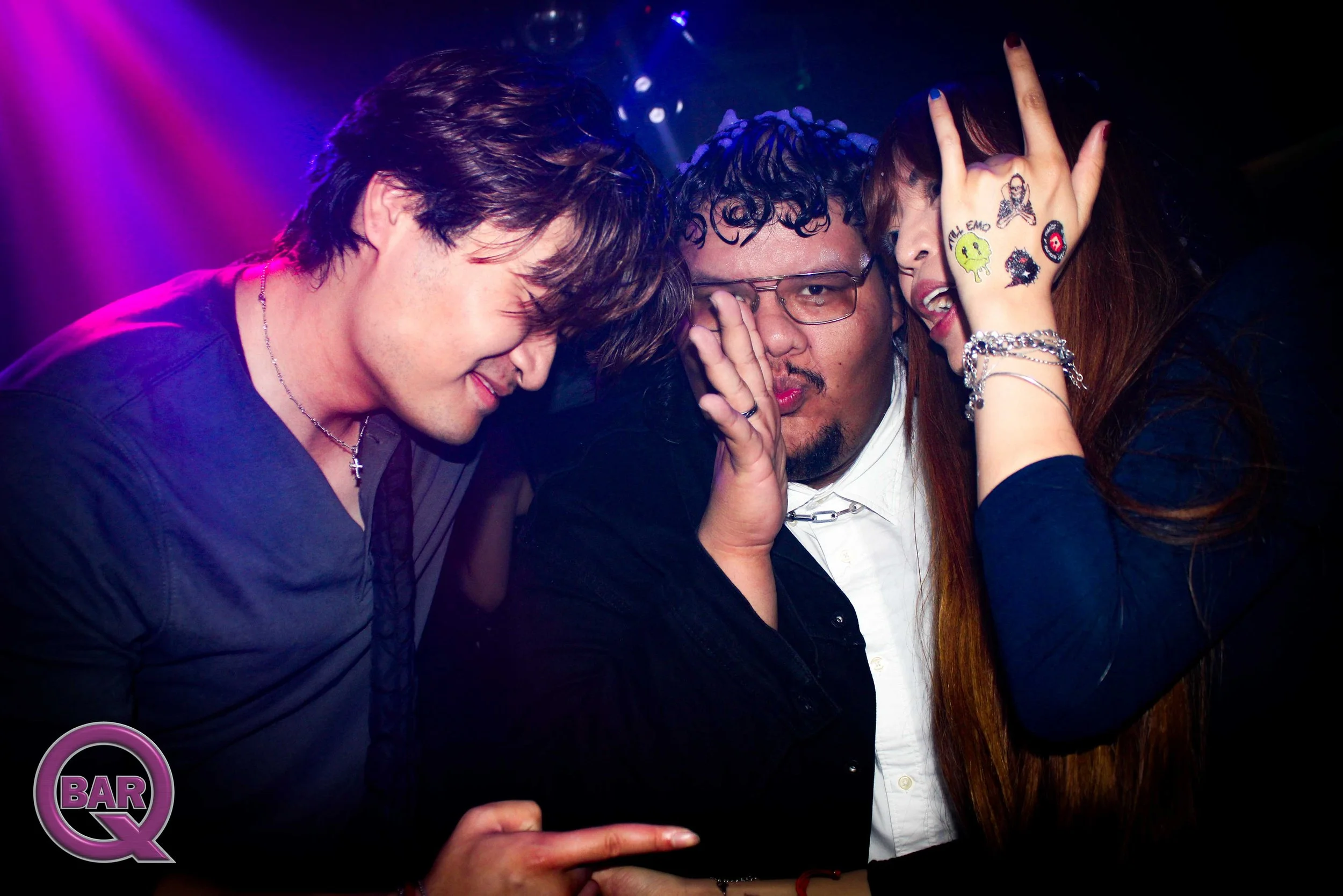 Three friends at a nightclub or party, two women and one man, with their heads close together, making funny faces, with one woman wearing tattoos on her hand and wearing jewelry, colorful lighting, and a disco ball in the background.