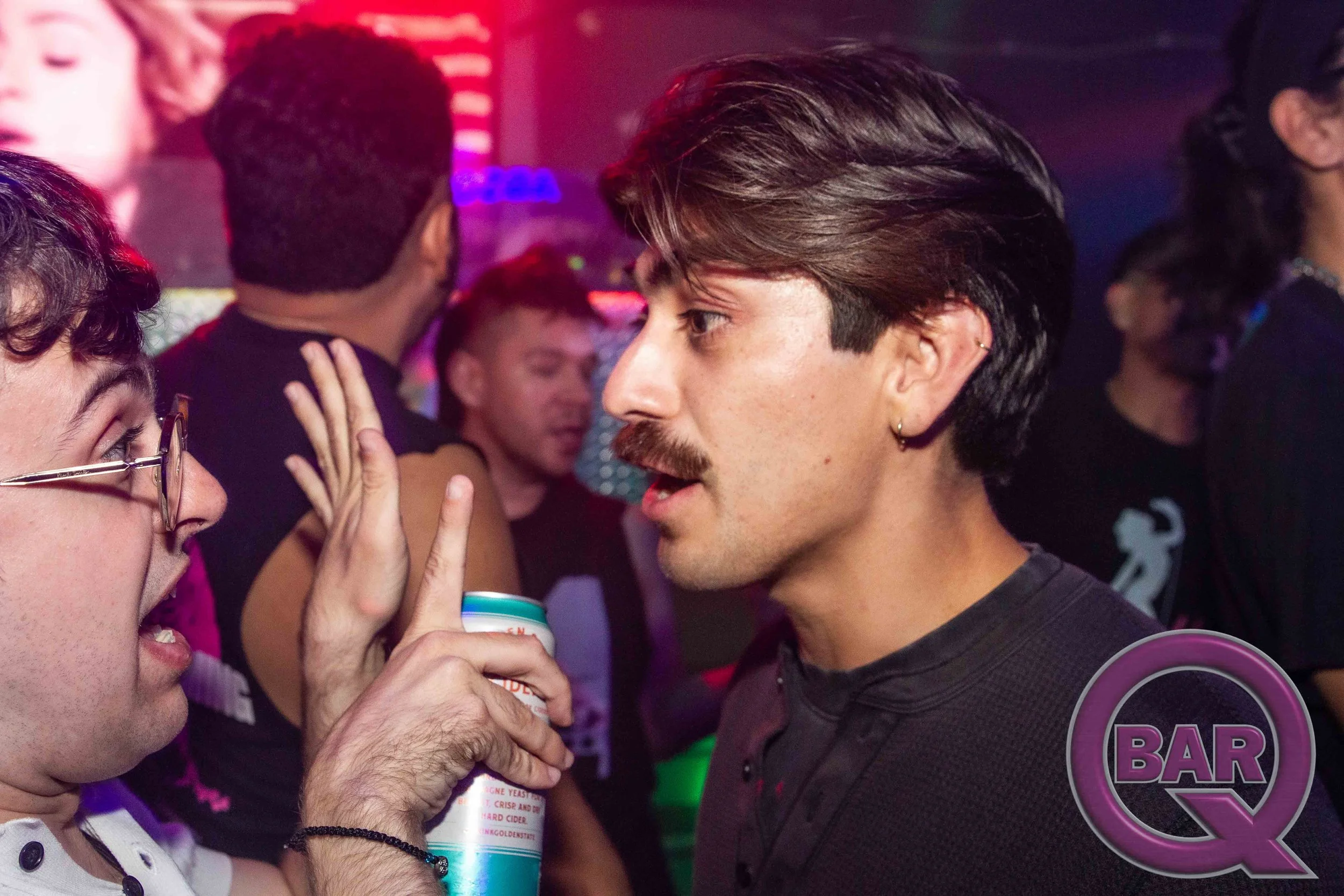 A woman with glasses and a man with dark hair and a mustache face each other closely. The woman is touching the man's cheek with her hand. The setting appears to be a bar or nightclub, with other people and colorful lighting in the background.